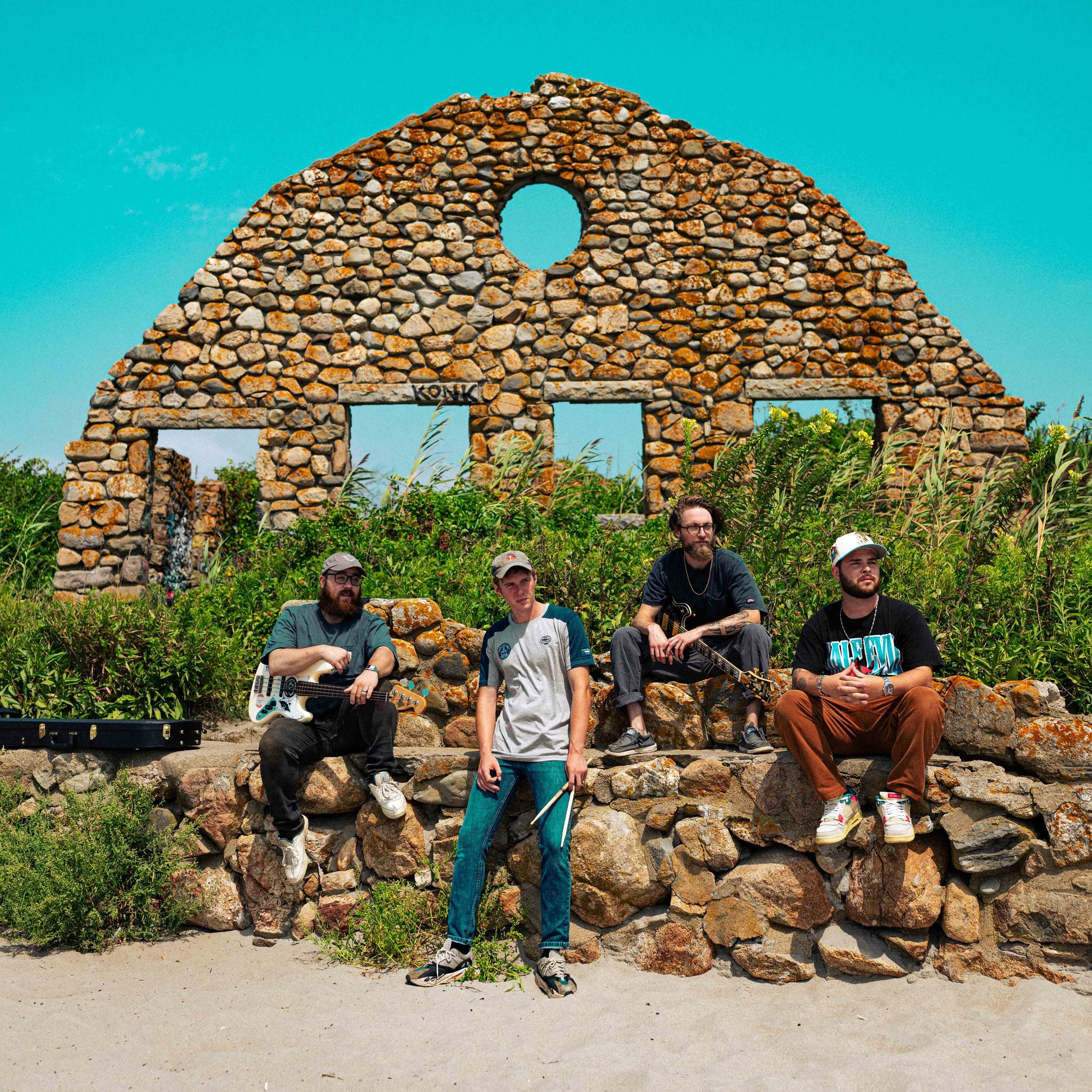 Four men with musical instruments sit and stand on a stone wall in front of a stone ruin with a circular window and two rectangular windows, surrounded by green bushes under a clear blue sky.