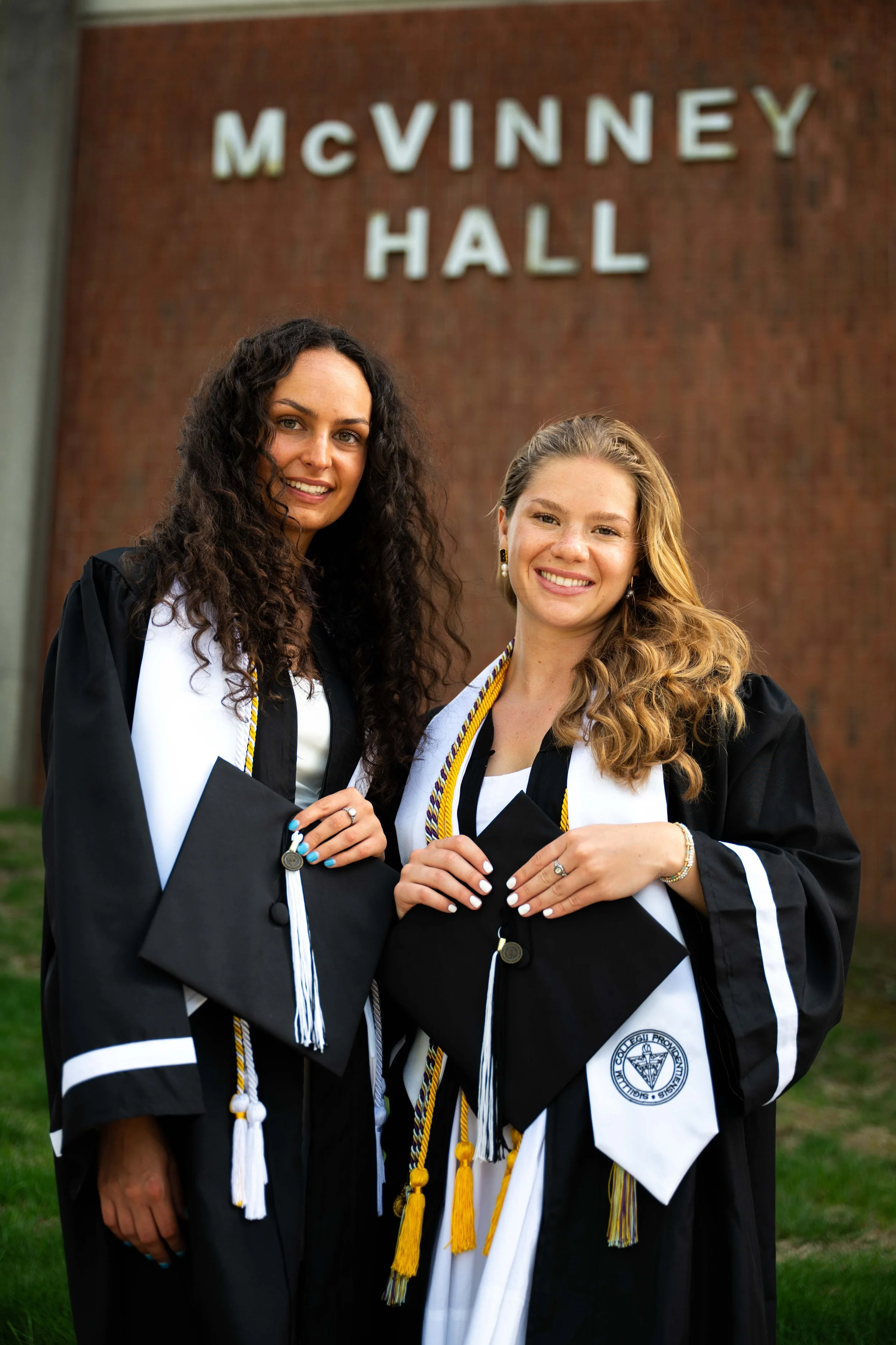 Two women in graduation gowns and caps standing in front of a brick building with a sign that reads "McVinney Hall."