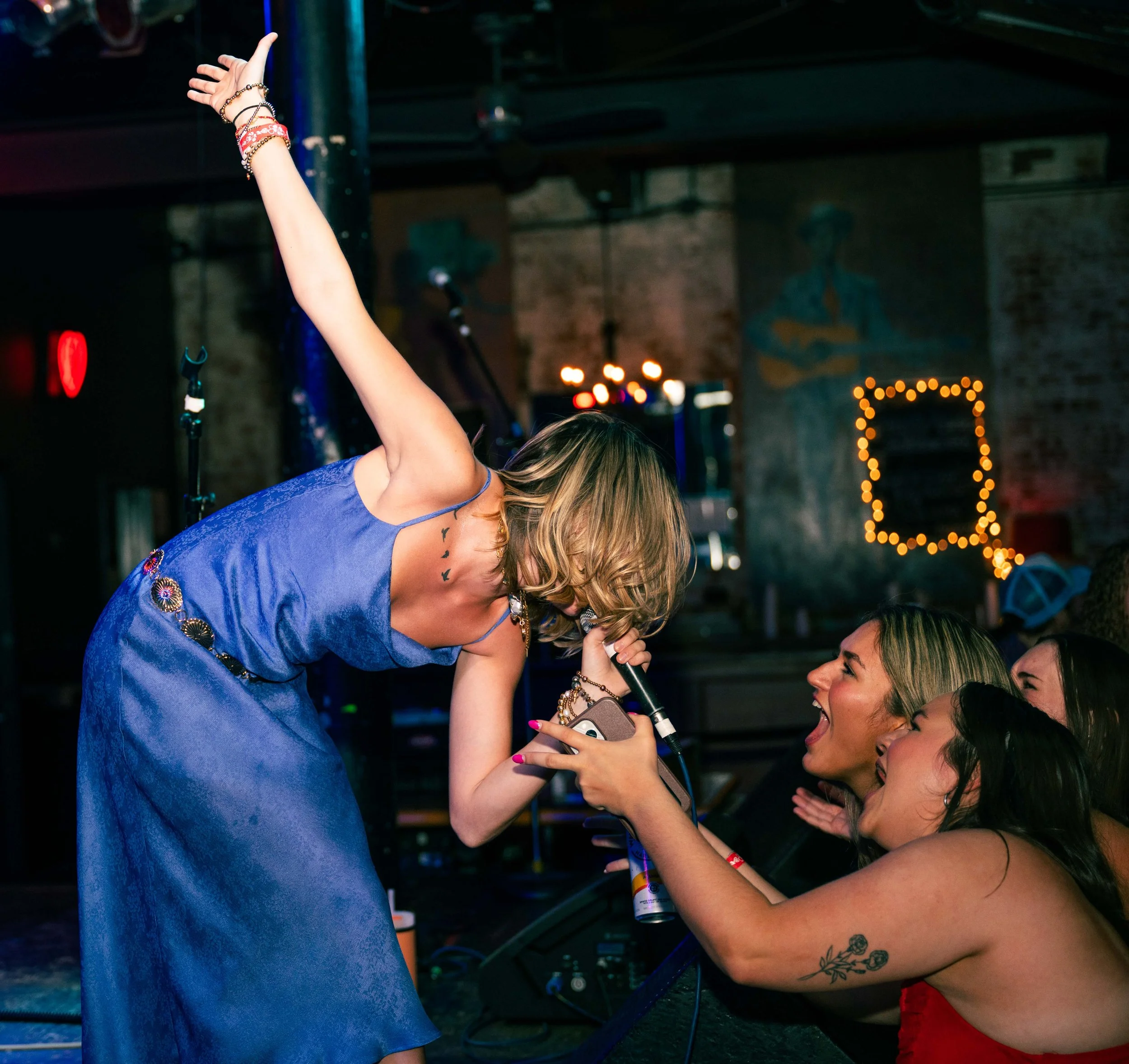 A woman performing on stage at a concert, leaning forward, singing into a microphone, wearing a blue dress. She interacts with the audience, who are reaching out towards her with excitement and smiles in a dimly lit venue with a mural and string ligh