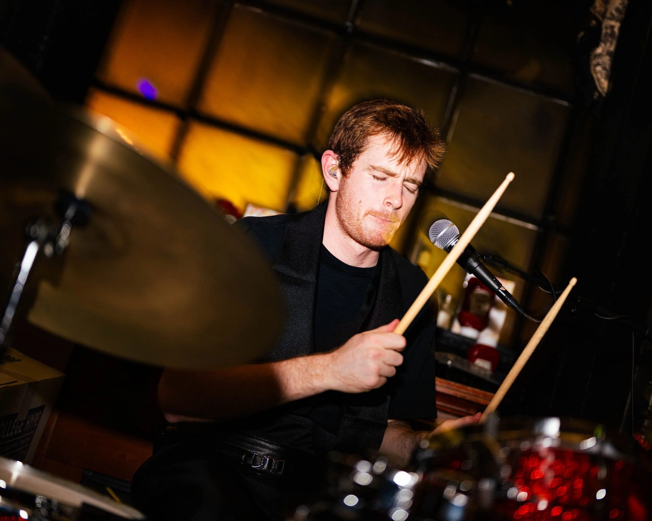 A young man with light skin, brown hair, and a focused expression playing drums in a dimly lit room, using drumsticks with a microphone nearby.