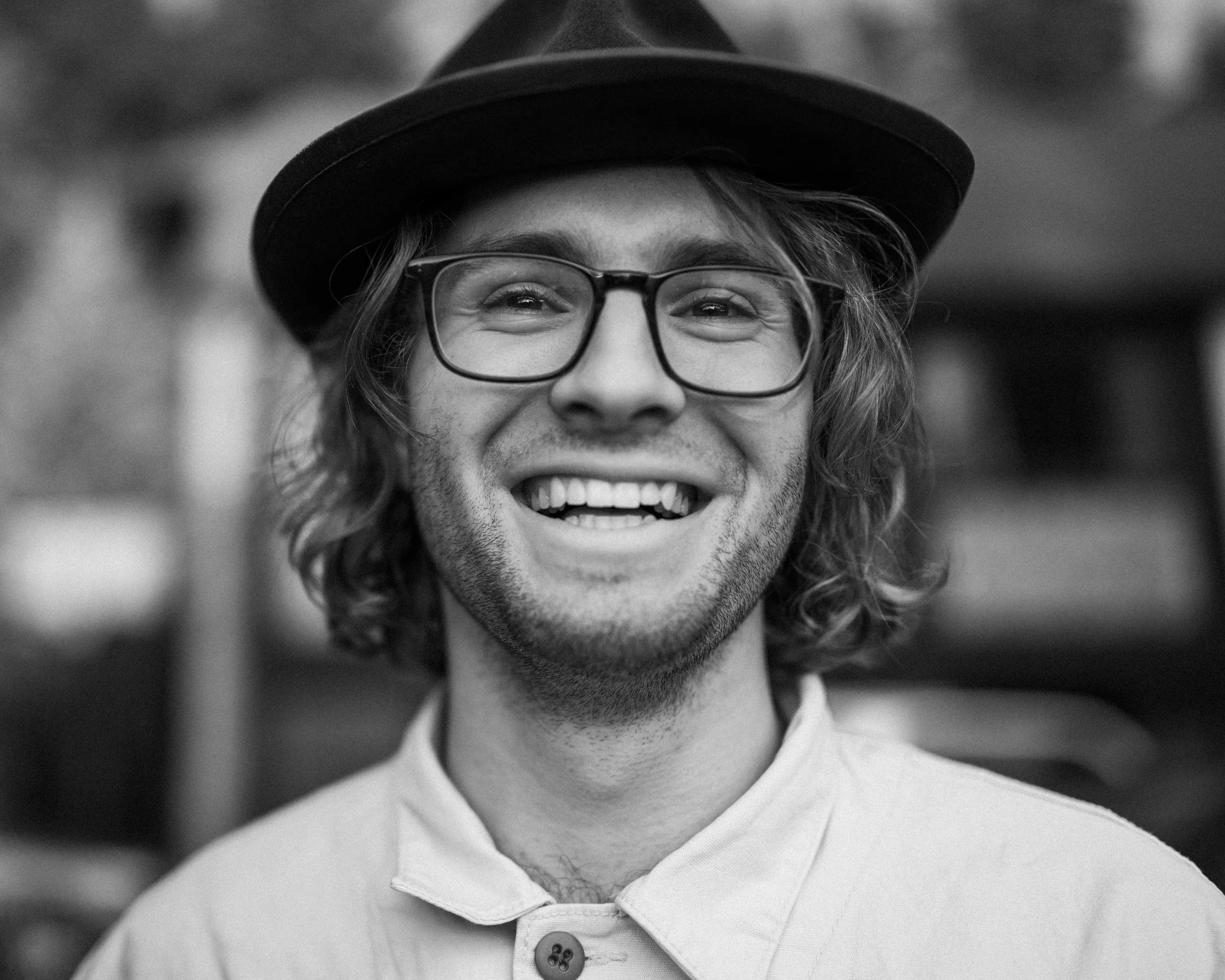Smiling young man with glasses, curly hair, wearing a hat and a collared shirt, in black and white.