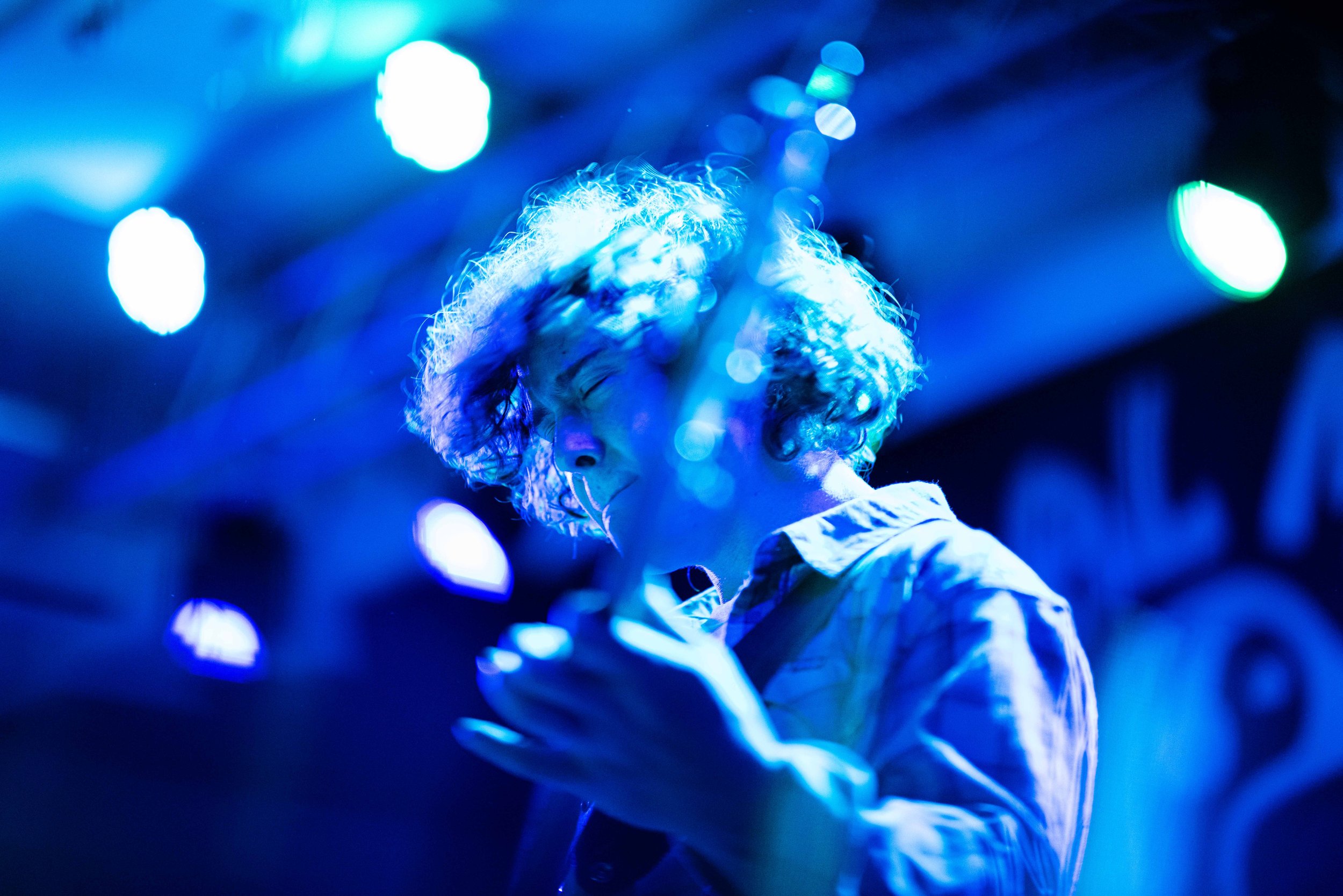 A person with curly hair illuminated by blue stage lighting, looking at their phone in a dimly lit environment, possibly at a concert or club.