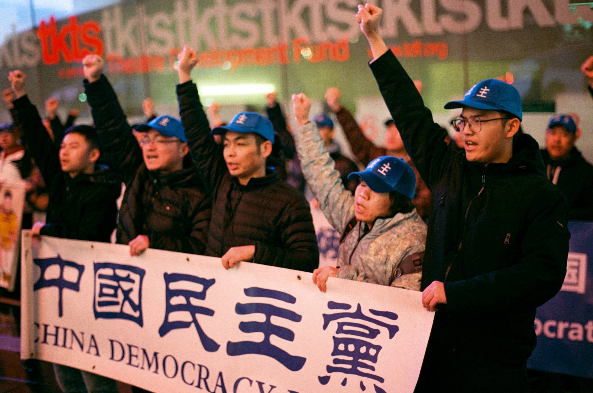 Group of protesters holding a banner with Chinese characters and the text 'China Democracy' in English in front of a building with a sign that reads '1kts'.