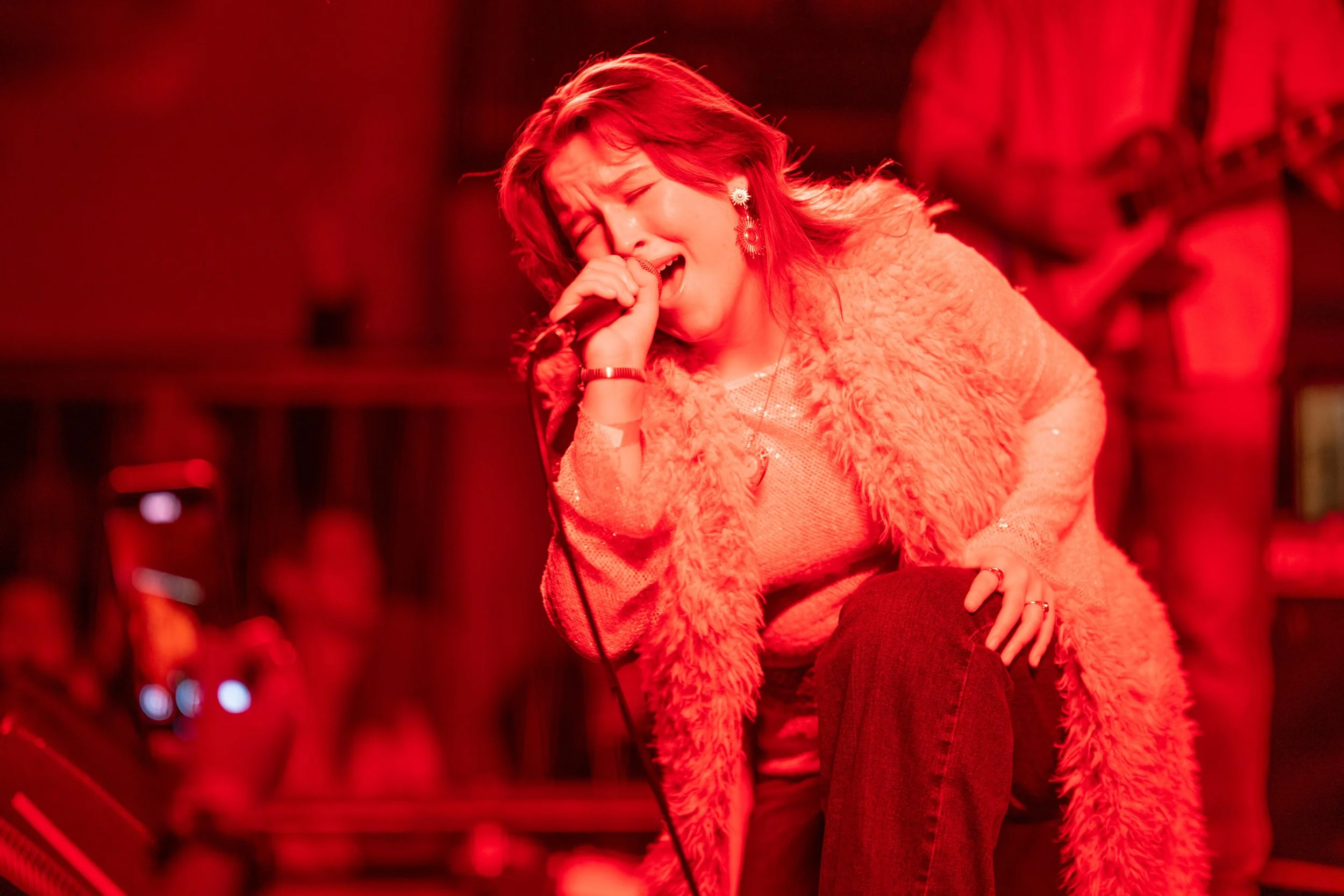A woman with red hair passionately singing into a microphone on stage, wearing a furry coat and jewelry, with a musician playing guitar in the background.