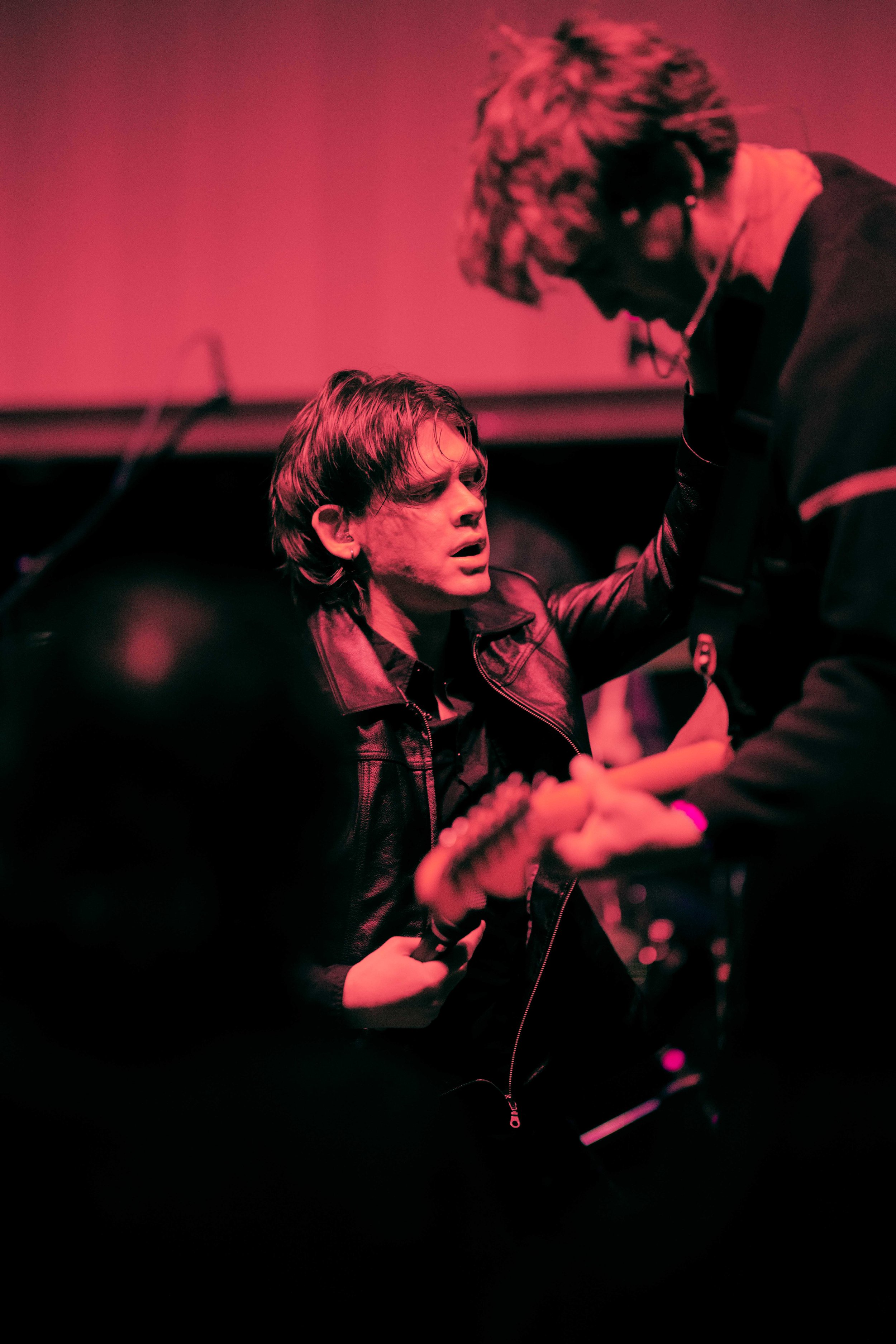 Two musicians performing on stage, one with a leather jacket playing a guitar, the other with curly hair and glasses leaning in, in a dimly lit setting with pink and black lighting.