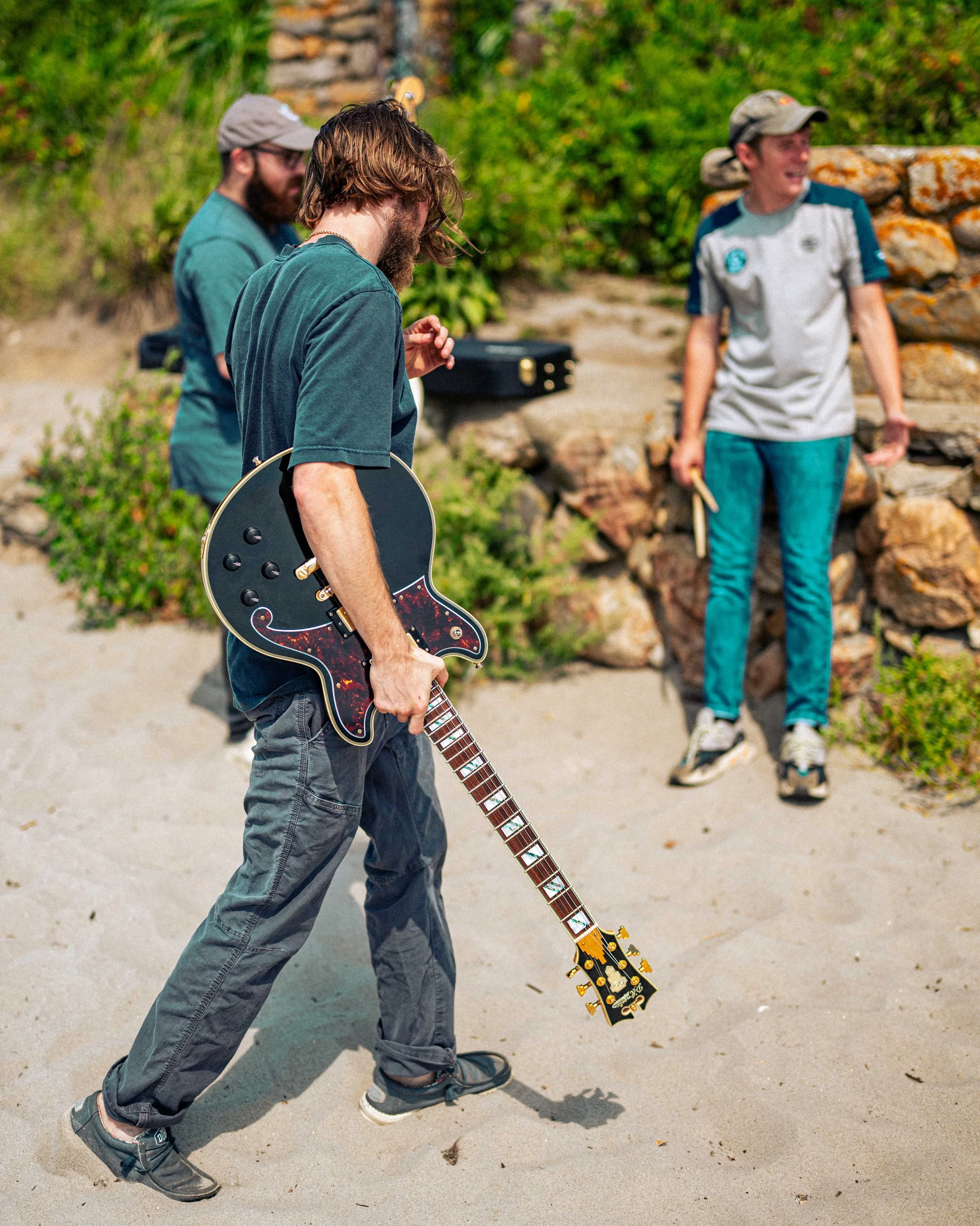 Three men are outdoors; one with long hair and glasses is holding an electric guitar, the second with a beard is wearing a cap with a beard, and the third is smiling and holding drumsticks.