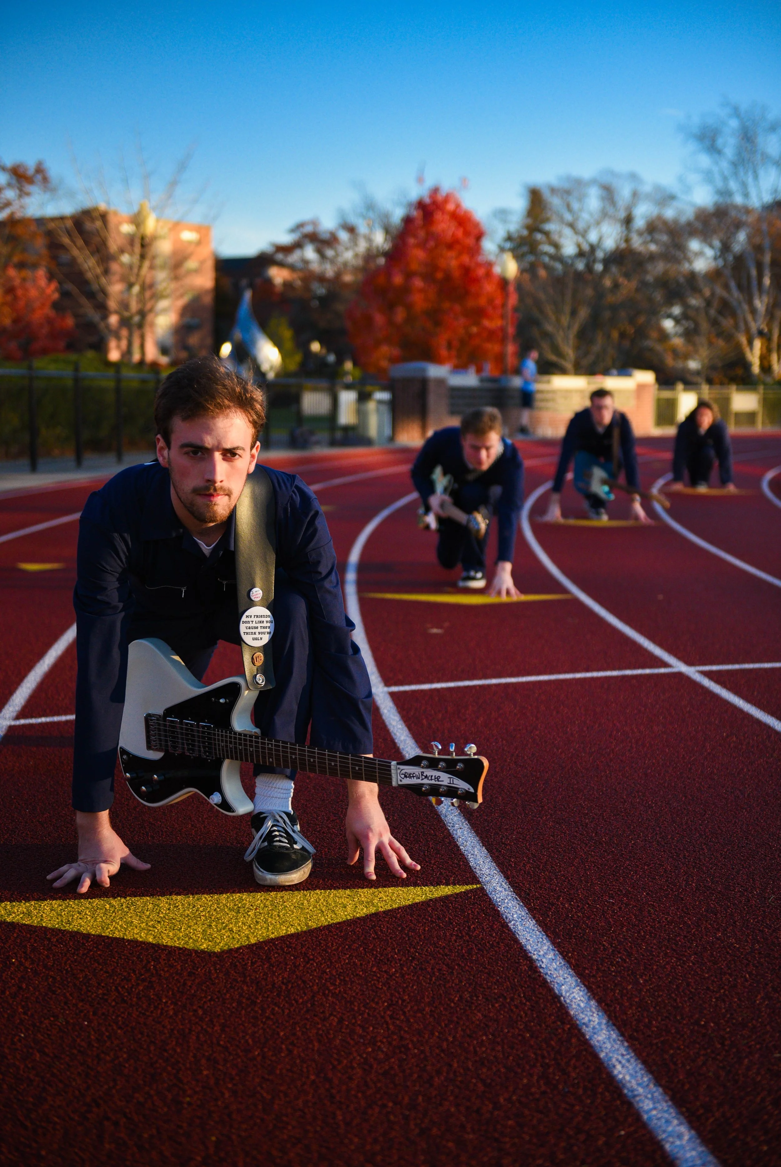 Young man with a guitar at starting line on a running track, with three other men behind him, preparing for a race, on a sunny day with trees and buildings in the background.