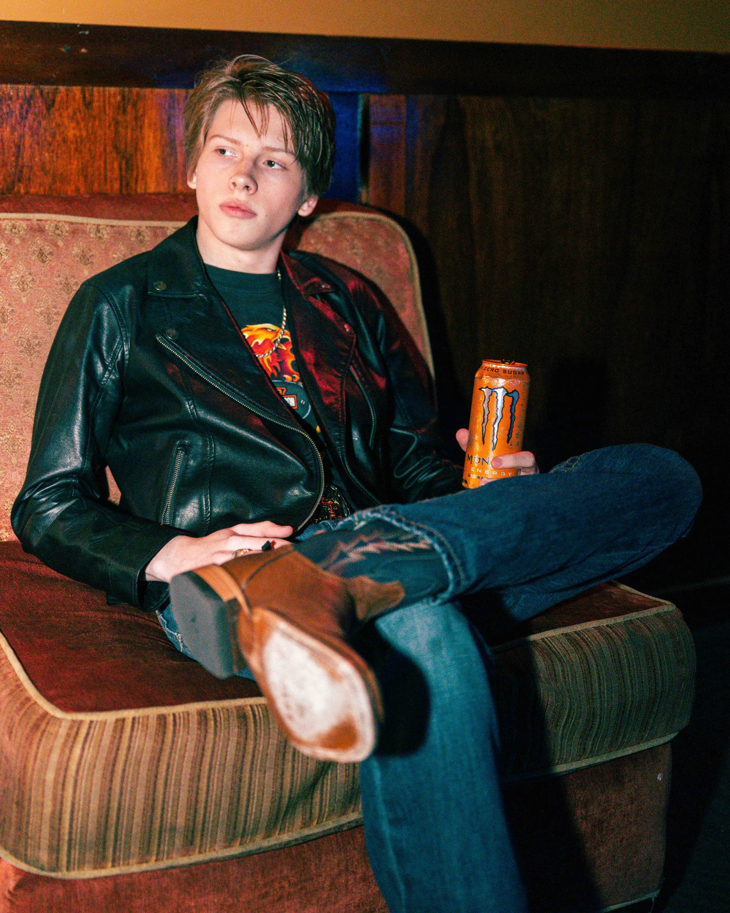 A young man with short hair sitting on a vintage style sofa, wearing a black leather jacket, jeans, and brown boots, holding an orange Monster energy drink can.