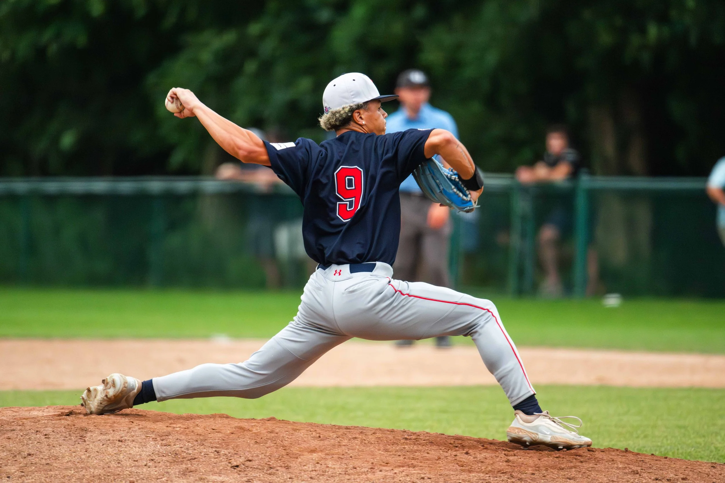 A baseball player in a dark blue jersey and white pants with red piping, wearing number 9, pitching on the baseball field.