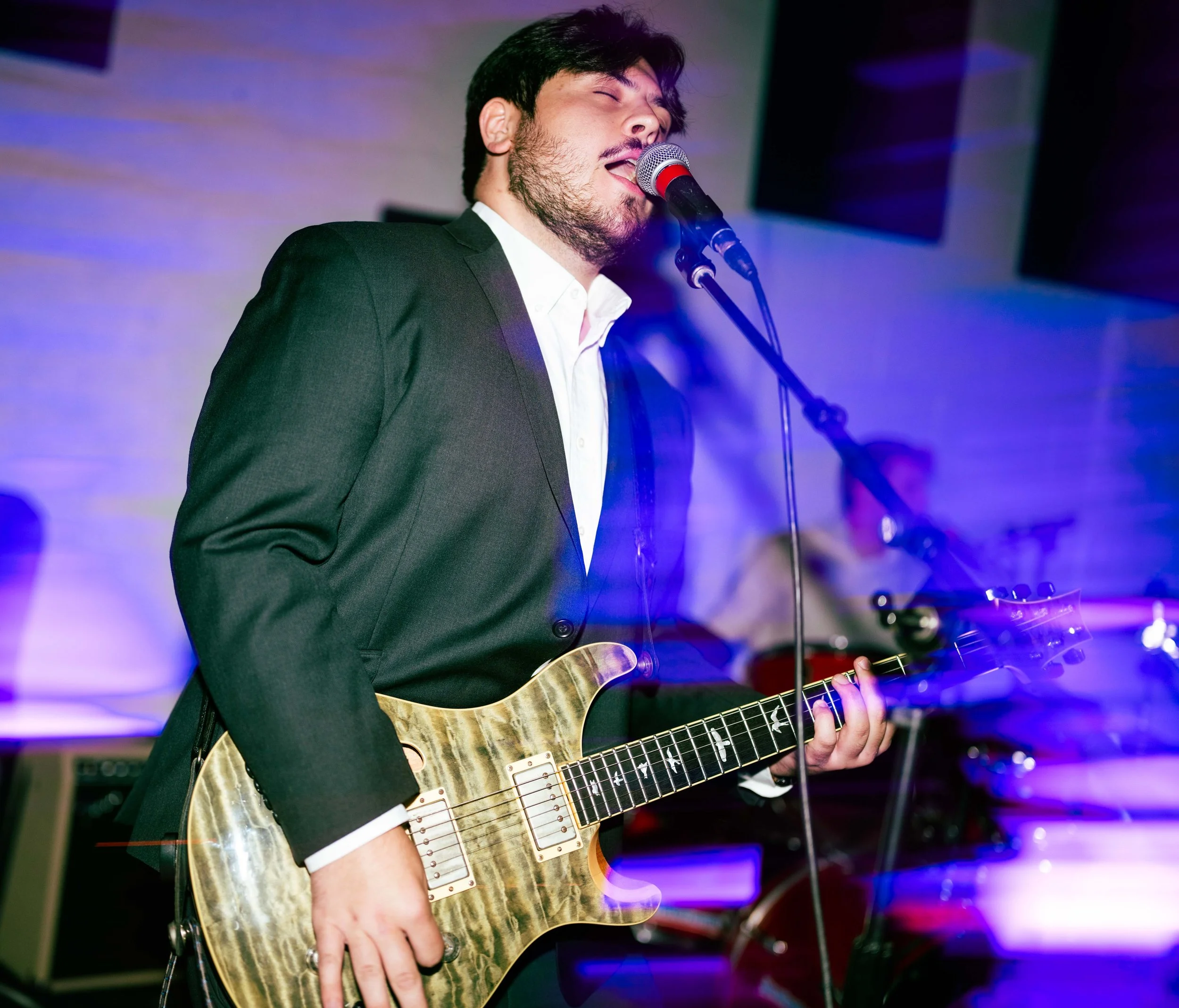 A man in a black suit and white shirt performs as a singer and guitarist on stage, singing into a microphone with his eyes closed and playing an electric guitar with a wood finish.