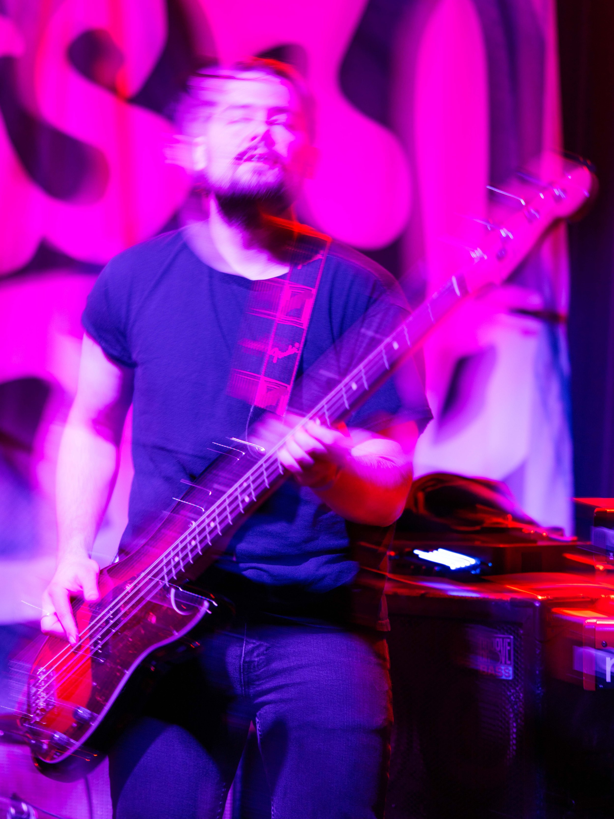 A musician playing an electric guitar on stage with motion blur effects due to movement, in front of a pink and purple background.