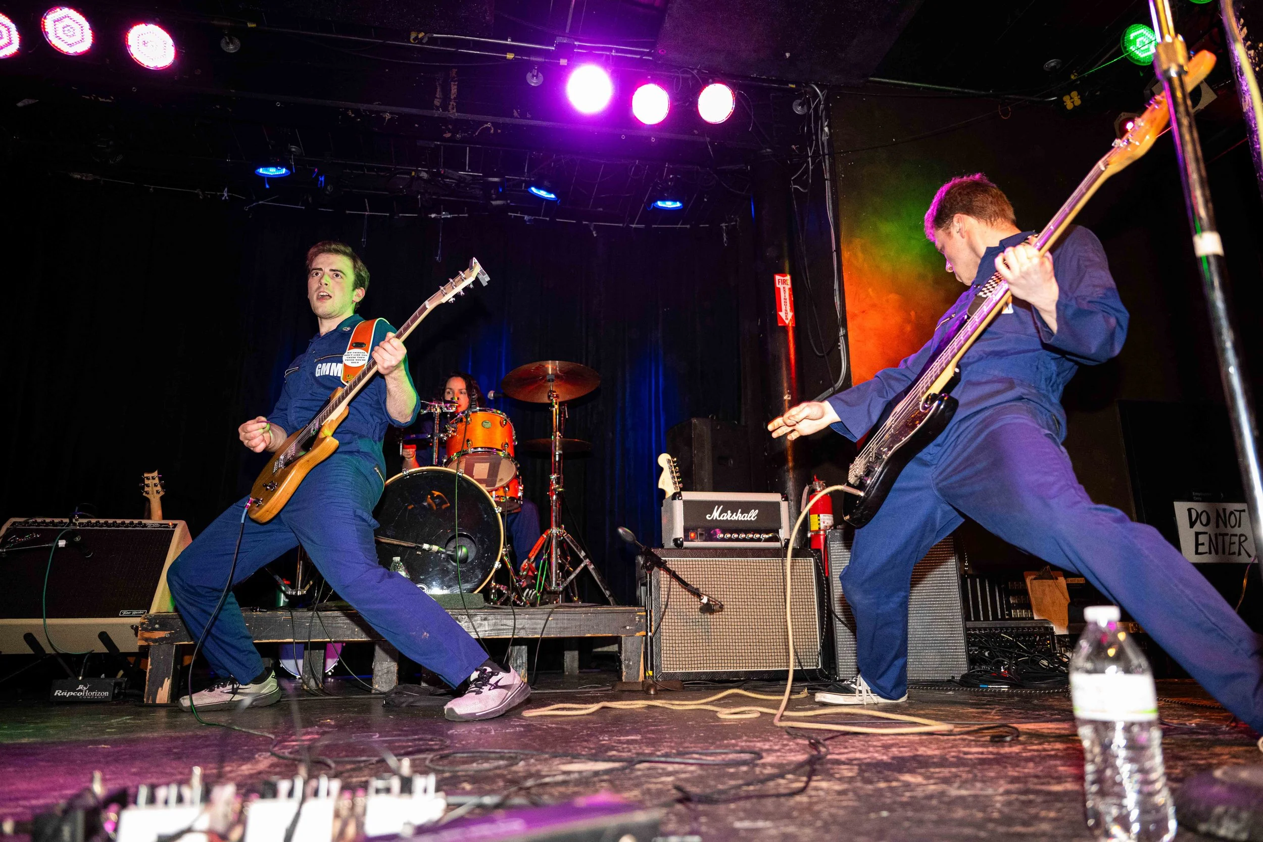 Two musicians playing electric guitars on stage with a drummer in the background, colorful stage lighting, and audio equipment in the foreground.
