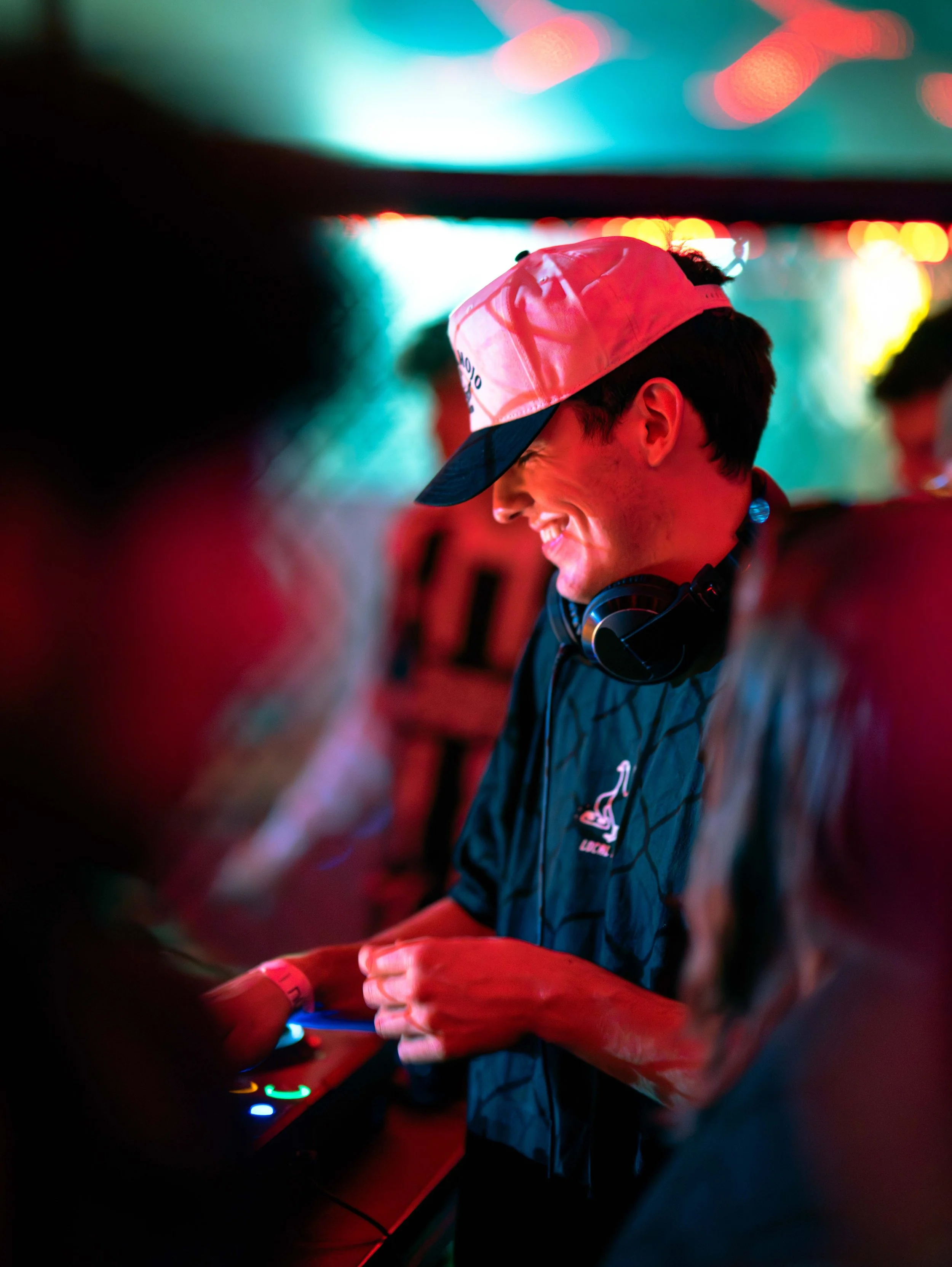 A young man with headphones, wearing a baseball cap, smiling while DJing at a colorful, vibrant party with neon lights.
