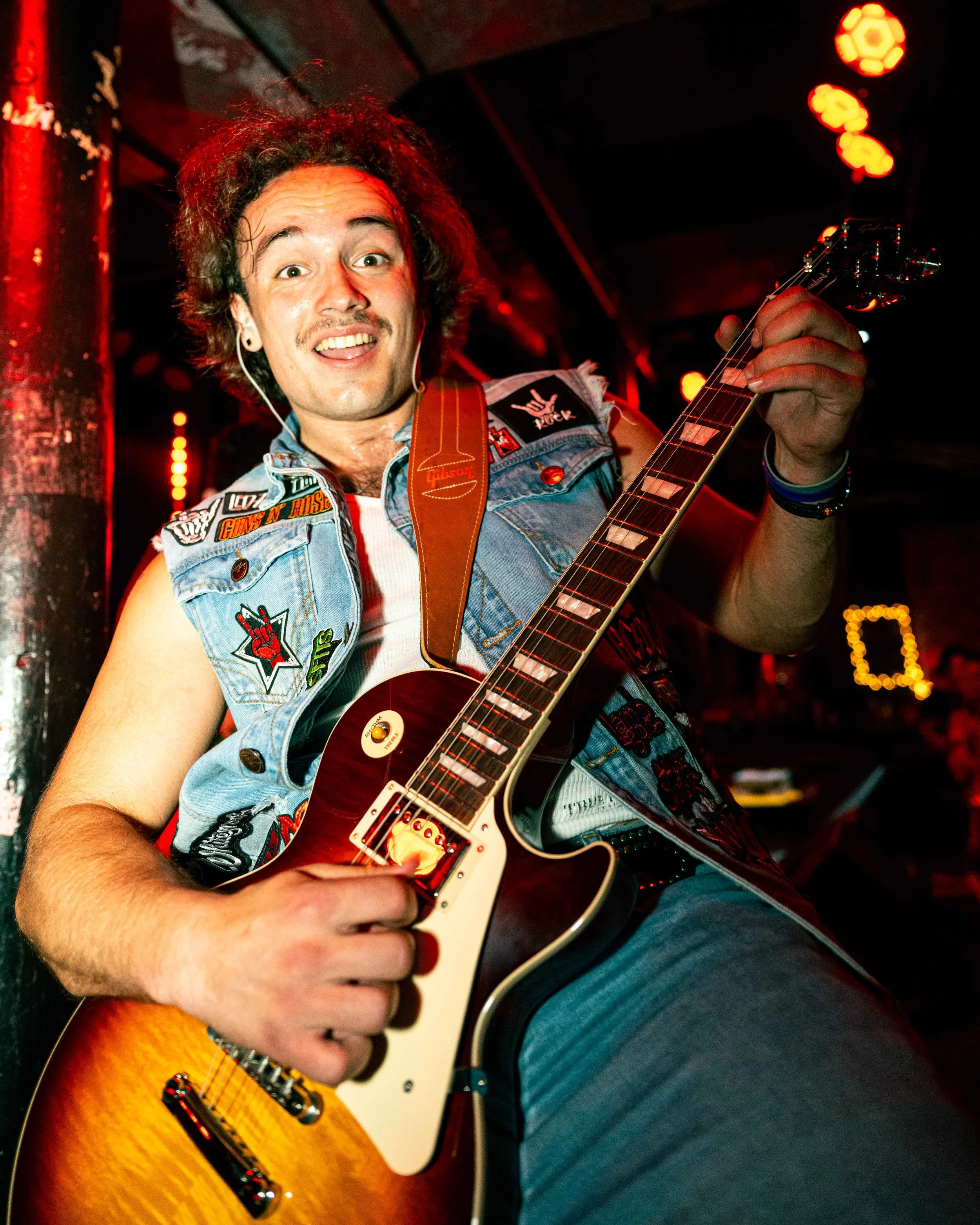 A musician playing an electric guitar in a dimly lit venue with red lighting, wearing a denim vest with patches and a white sleeveless shirt, looking excited.