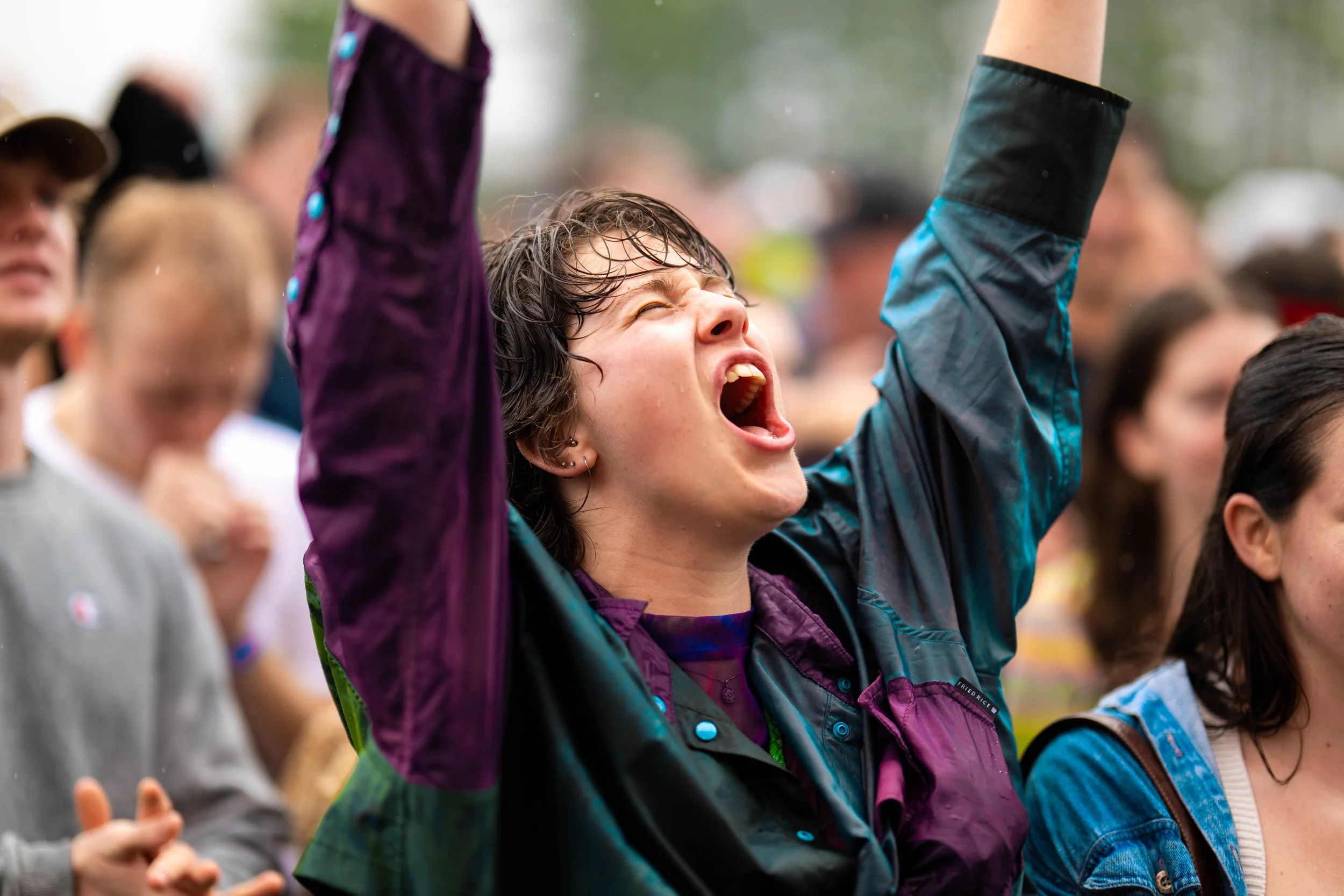 A woman with wet hair raising her arms and shouting passionately at a crowd during an outdoor event.