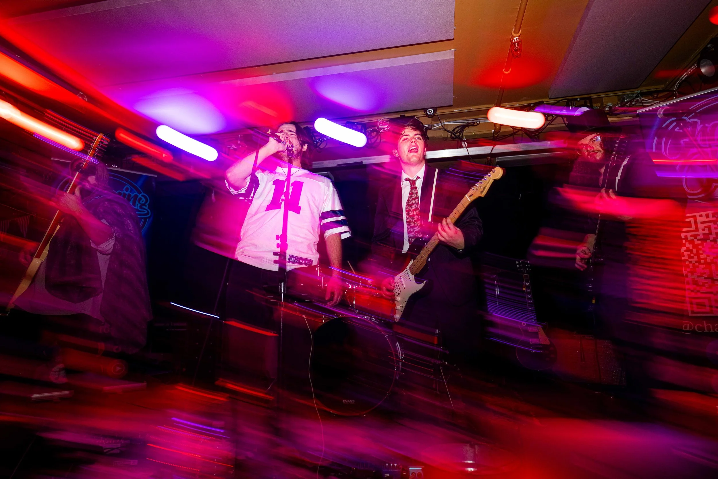 Band performing live on stage with colorful lighting, including a singer, guitarist, and a drummer