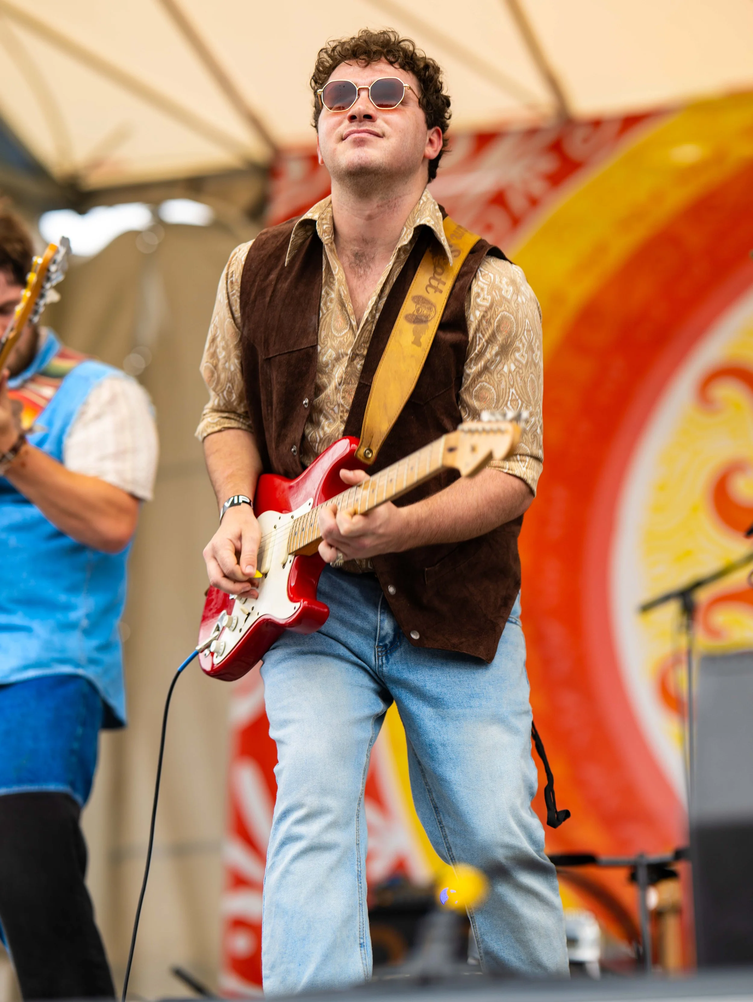 A young man wearing sunglasses, a patterned shirt, and a brown vest playing a red electric guitar on stage during a live music performance. Part of another musician playing a guitar is visible on the left, with a colorful backdrop behind them.