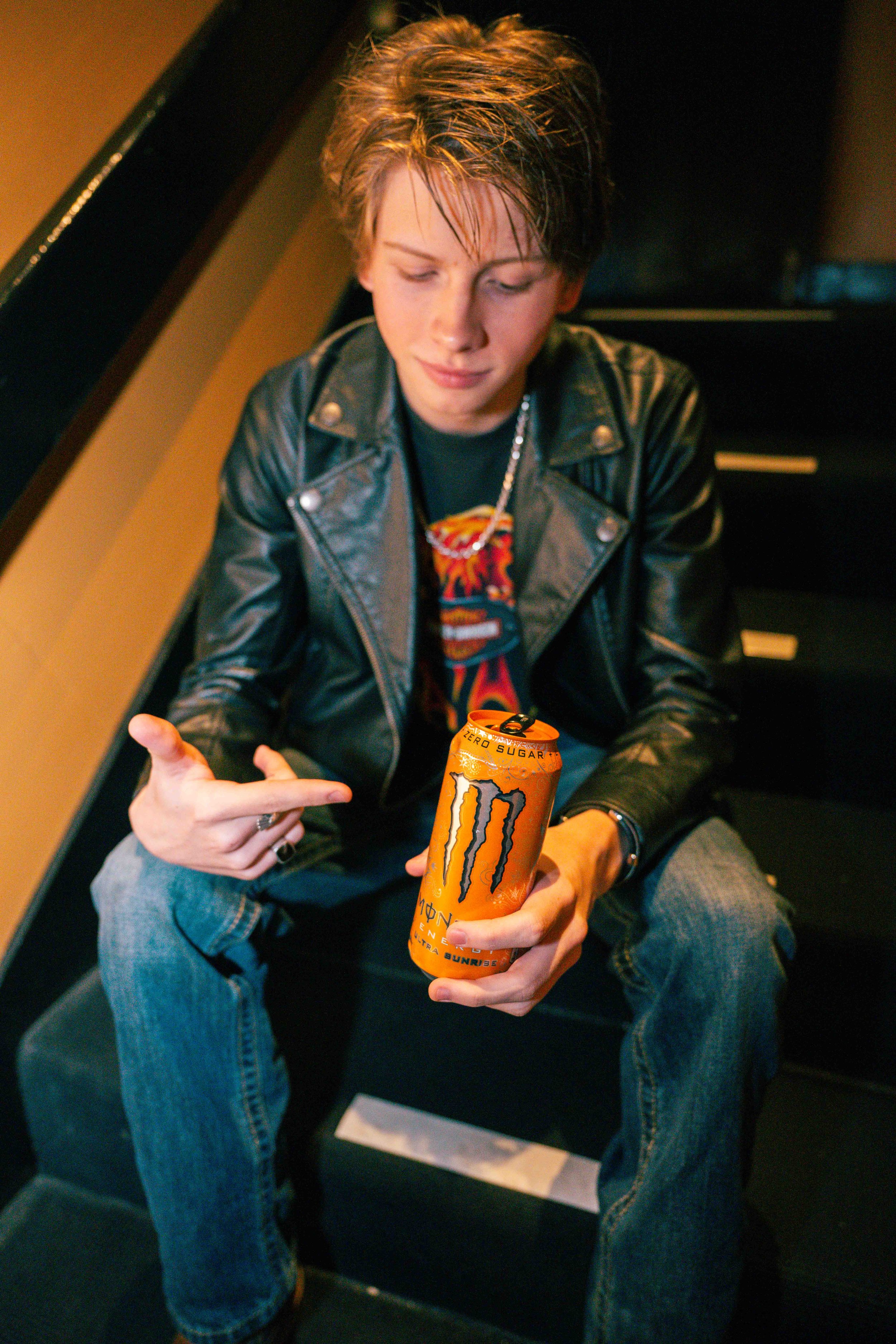 Young man with smudged makeup or face paint, wearing a leather jacket, sitting on a black bench, holding an orange Monster energy drink, pointing at it with his index finger, with tousled hair and a slight smile.