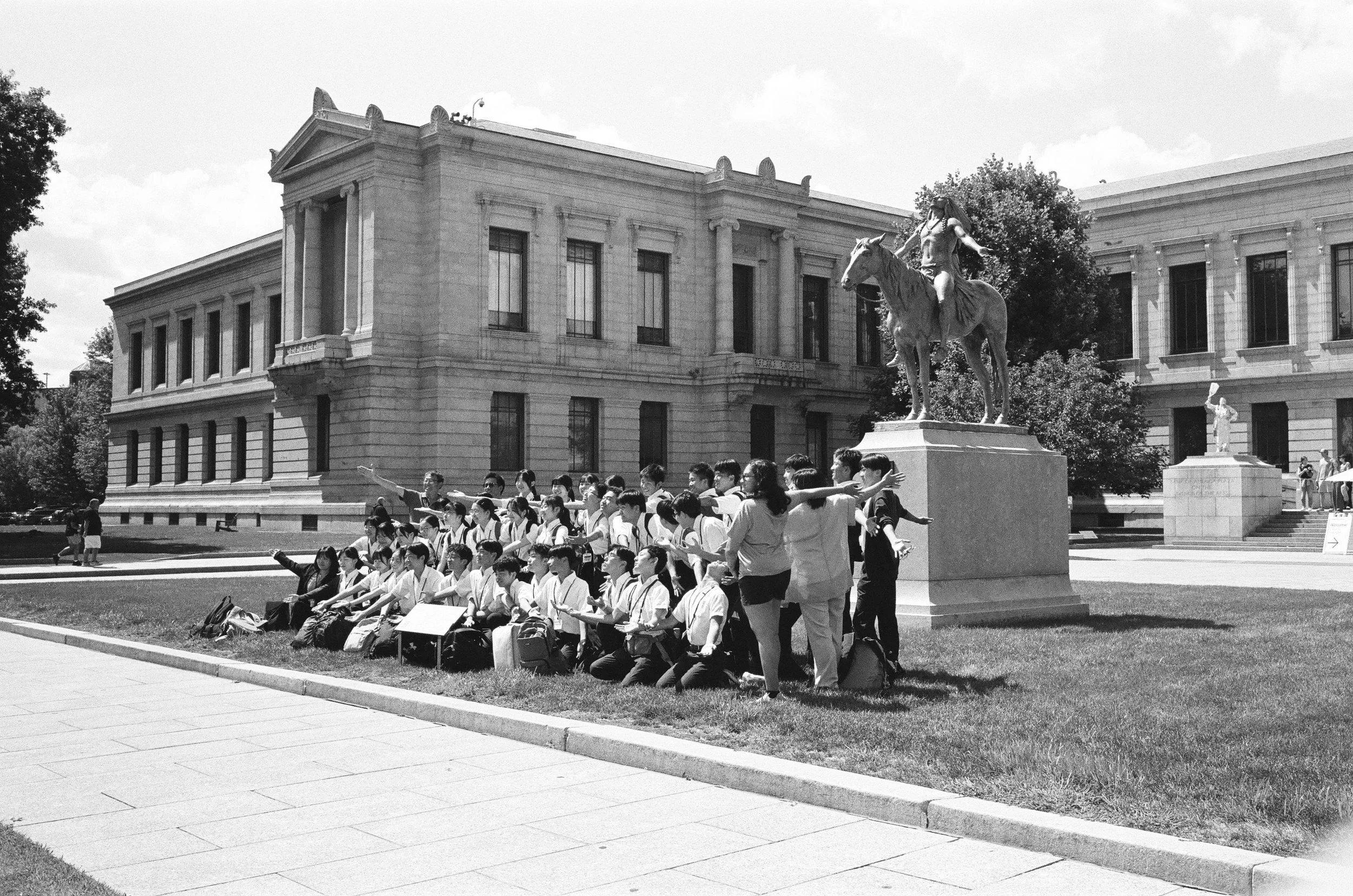 A group of students and teachers taking a tour outside a historic building with statues, including one of a rider on a horse, on a sunny day.