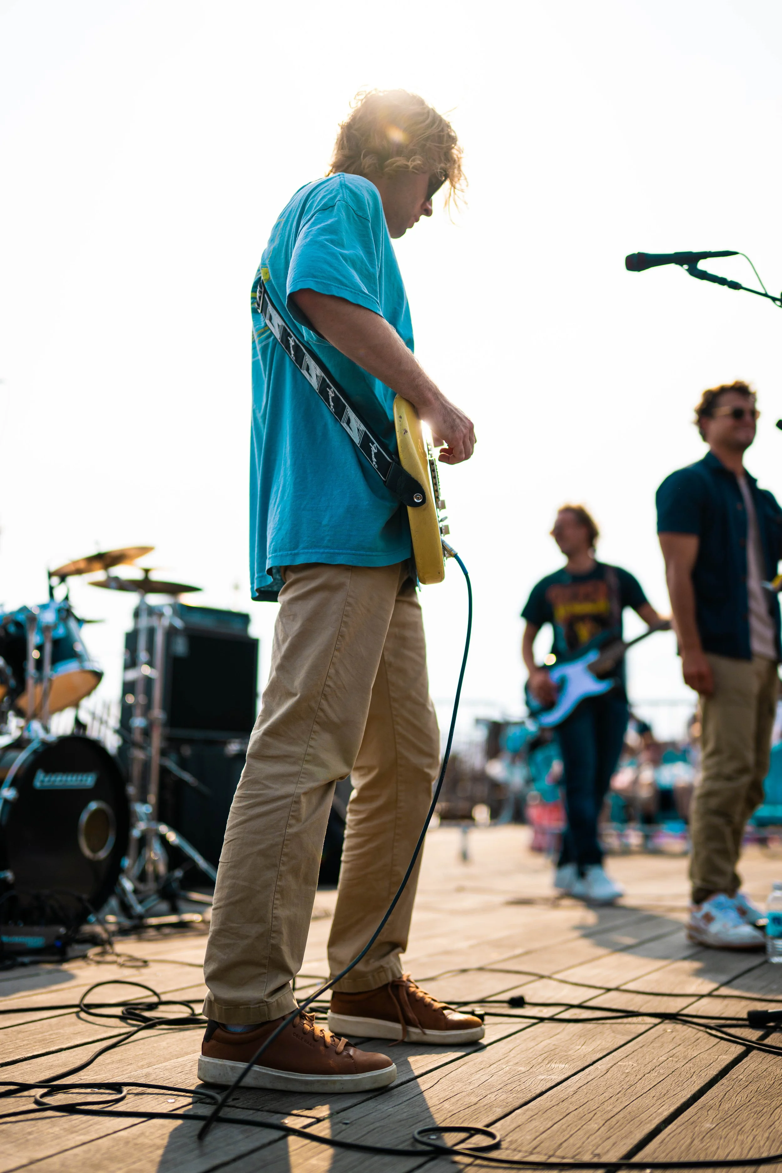 A person playing an electric guitar on stage with a band during an outdoor performance, with bright sunlight creating a silhouette effect.
