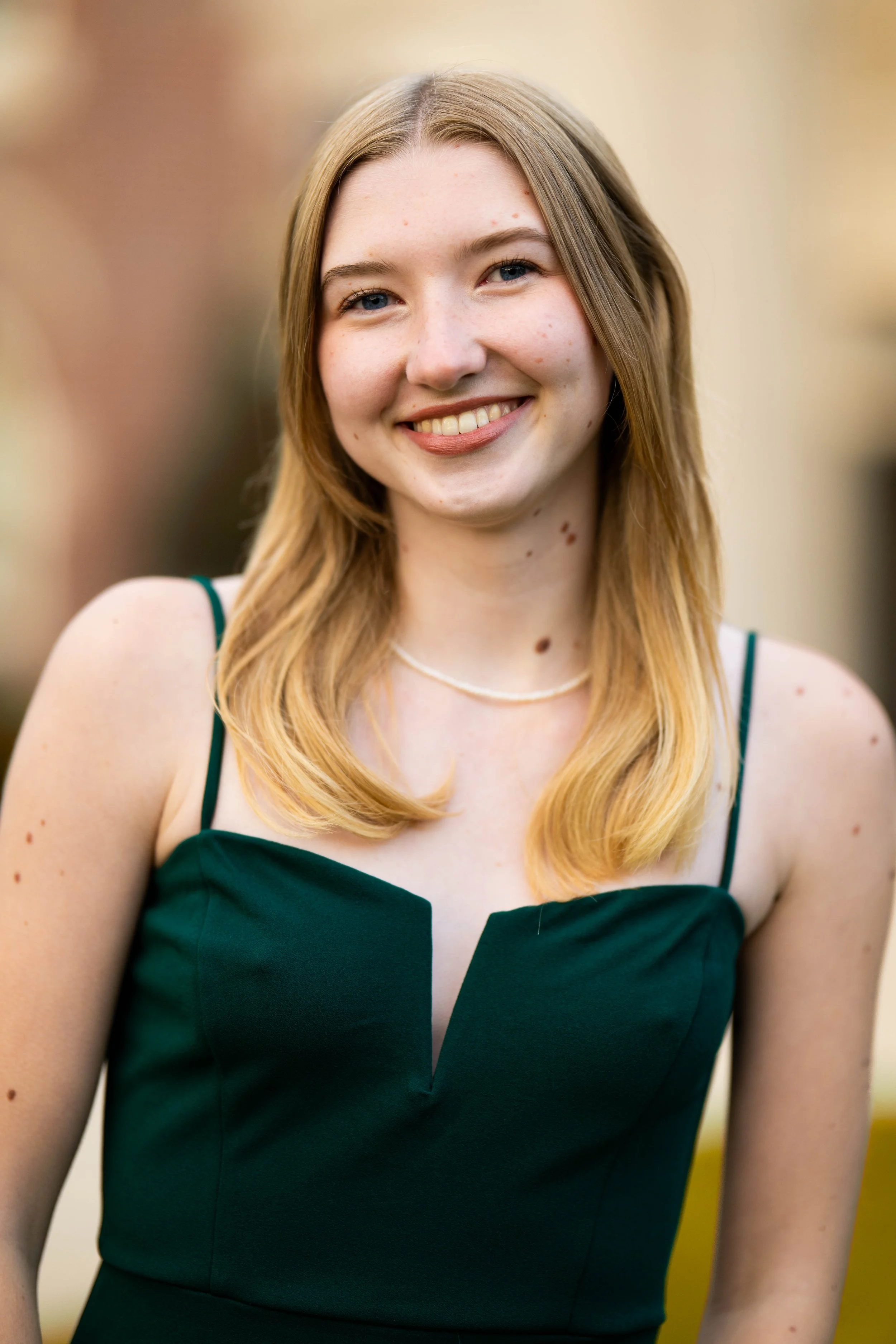 A young woman with blonde hair wearing a dark green dress with thin straps and a white necklace, smiling outdoors.