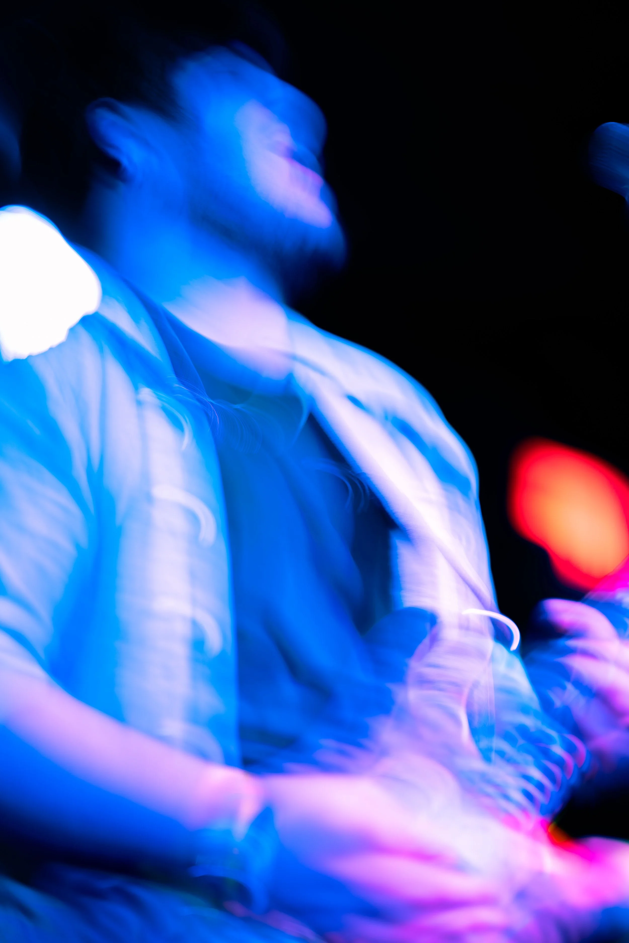 Blurred image of a person with glowing blue and red lights, wearing a white shirt, looking to the side, in a dark environment.