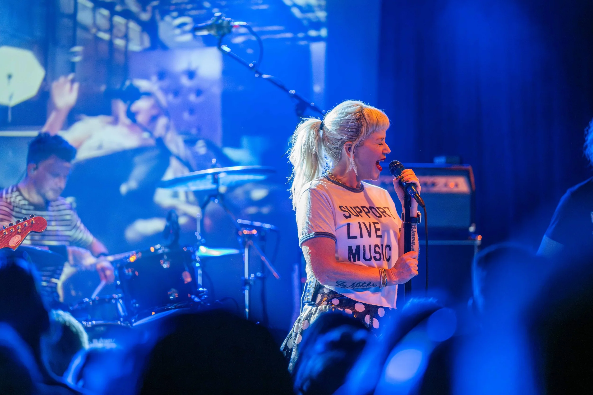 A woman with blonde hair in a ponytail singing into a microphone during a live music performance, wearing a white T-shirt with 'Support Live Music' written on it, and a patterned skirt, with band members and instruments visible in the background unde