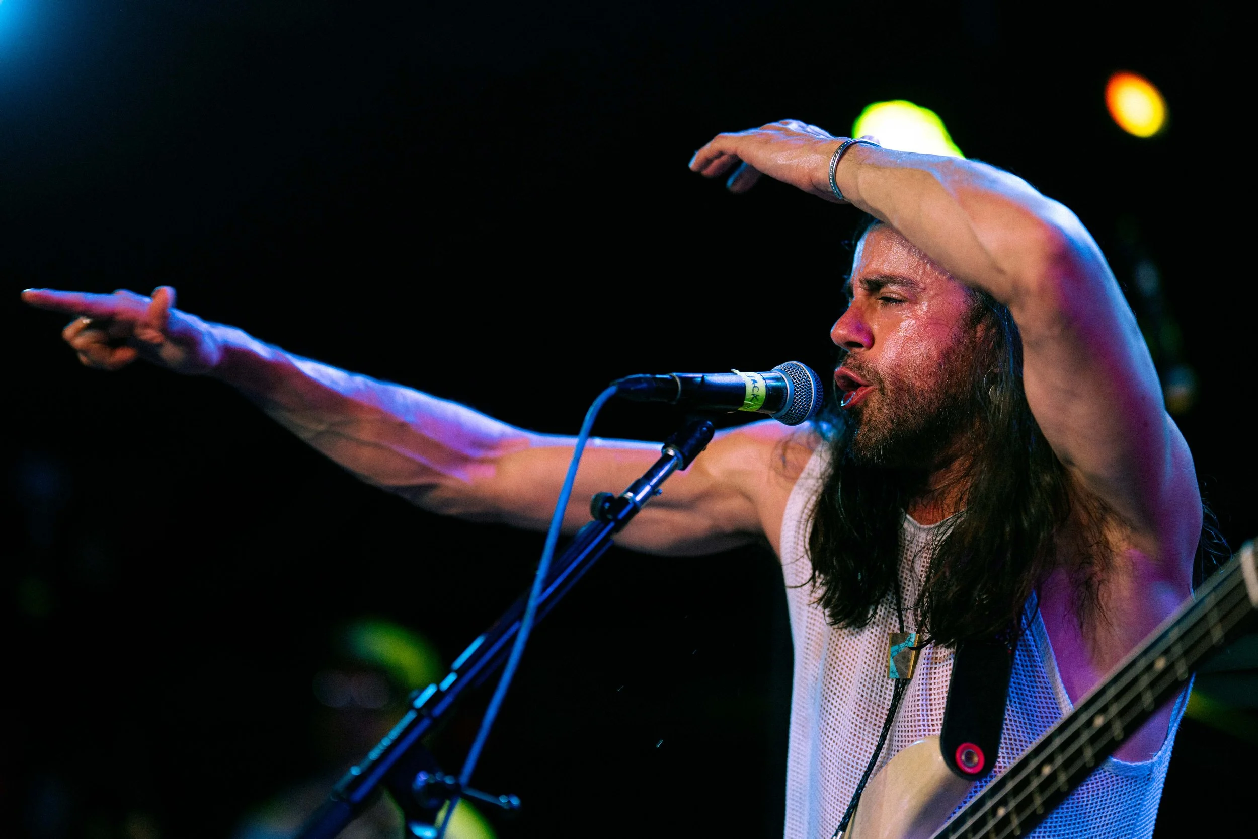 Musician with long hair and beard singing into a microphone while playing guitar, wearing a sleeveless shirt, with his arm extended outward.