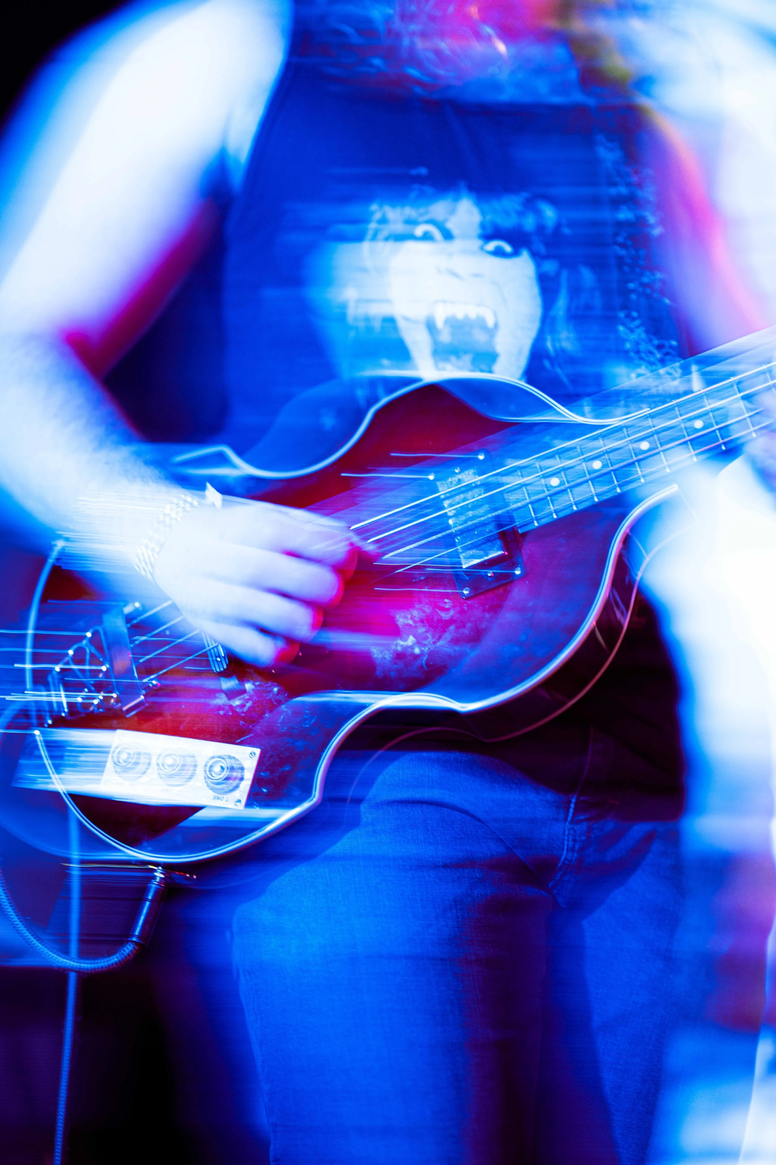 Person playing an electric guitar, wearing a t-shirt with a screaming face, with colorful blue and pink lighting effects.