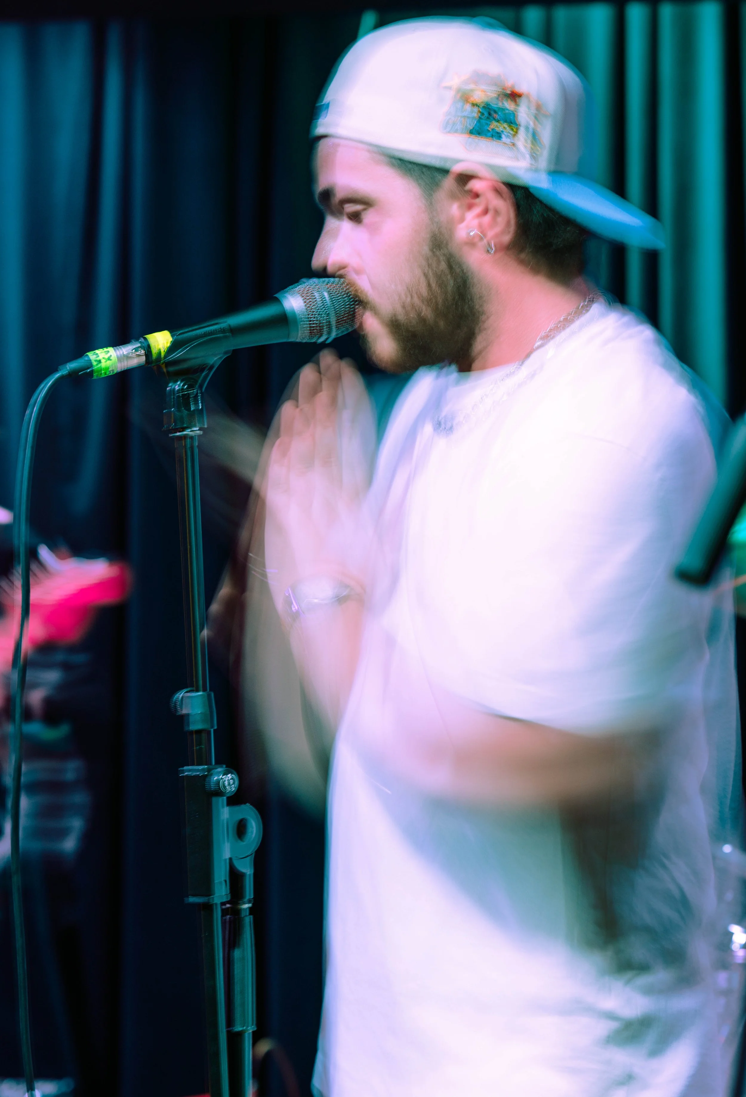 A male singer with a beard, wearing a white t-shirt and a white baseball cap backwards, performs with a microphone on a stage with dark curtains in the background.