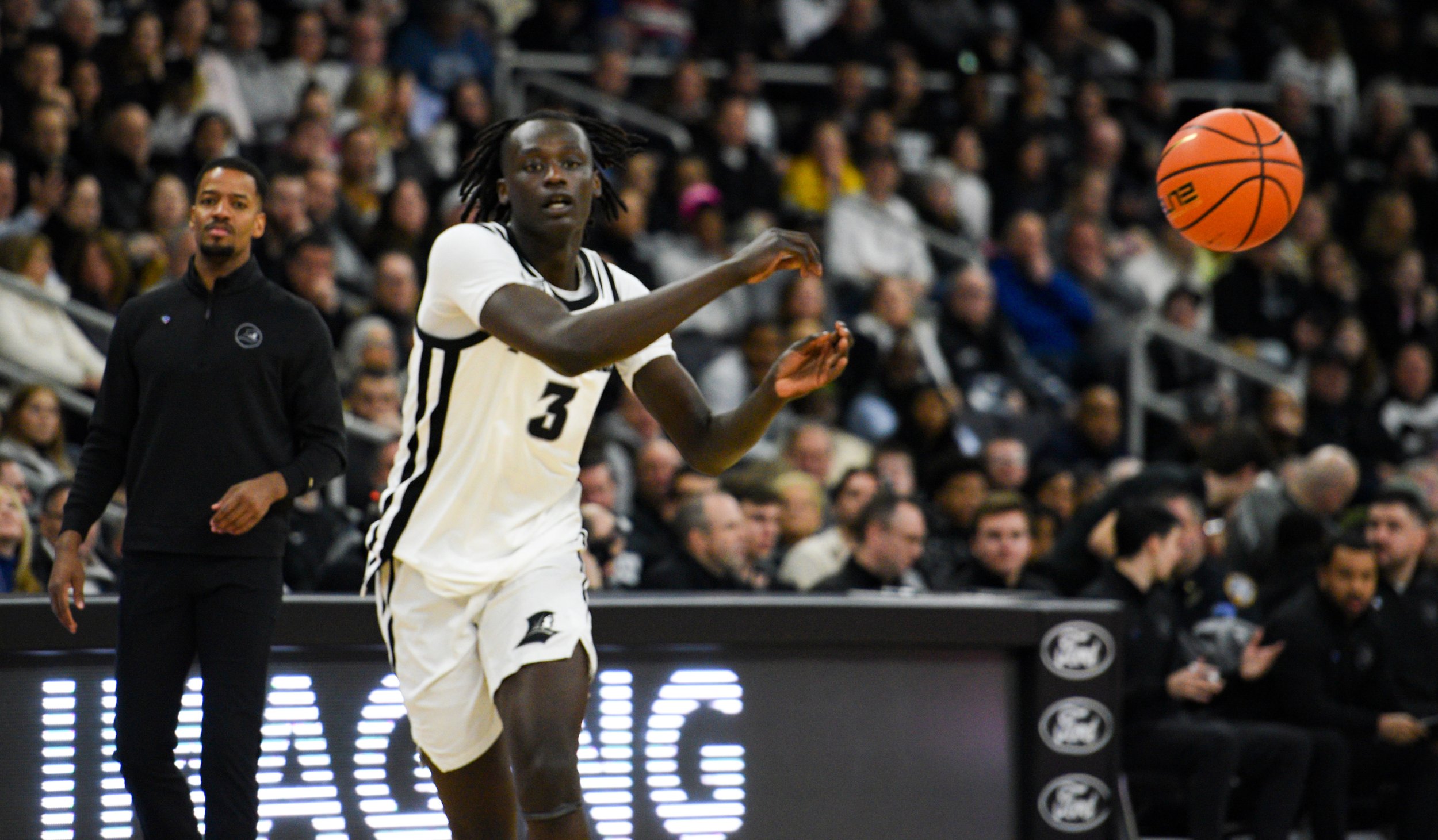 A basketball player in a white jersey with the number 3 is shooting a basketball during a game, with a crowd of spectators and a coach or referee in the background.