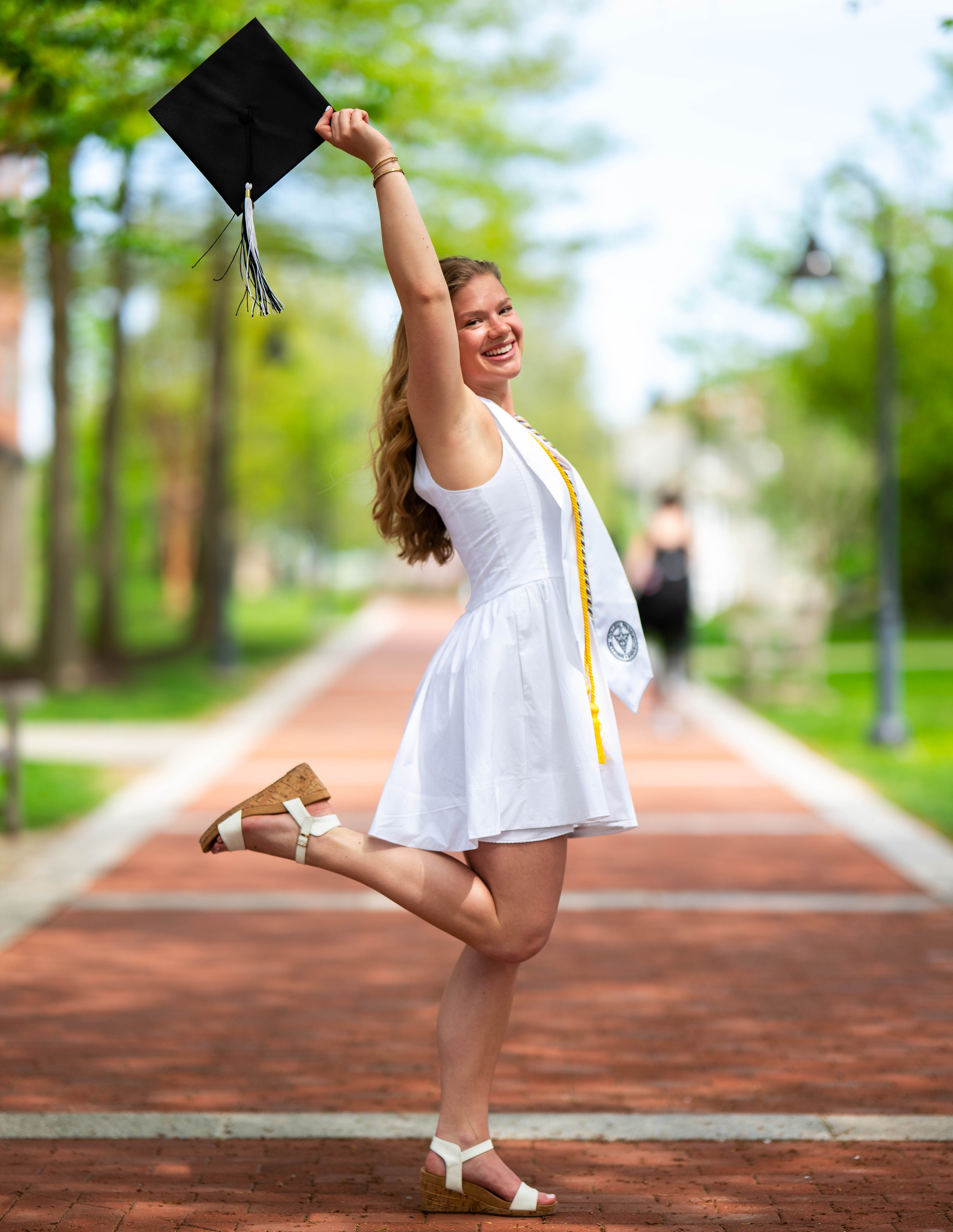 A young woman in a white graduation dress celebrating outdoors on a brick pathway, holding her graduation cap up in the air, smiling and posing with one foot lifted.
