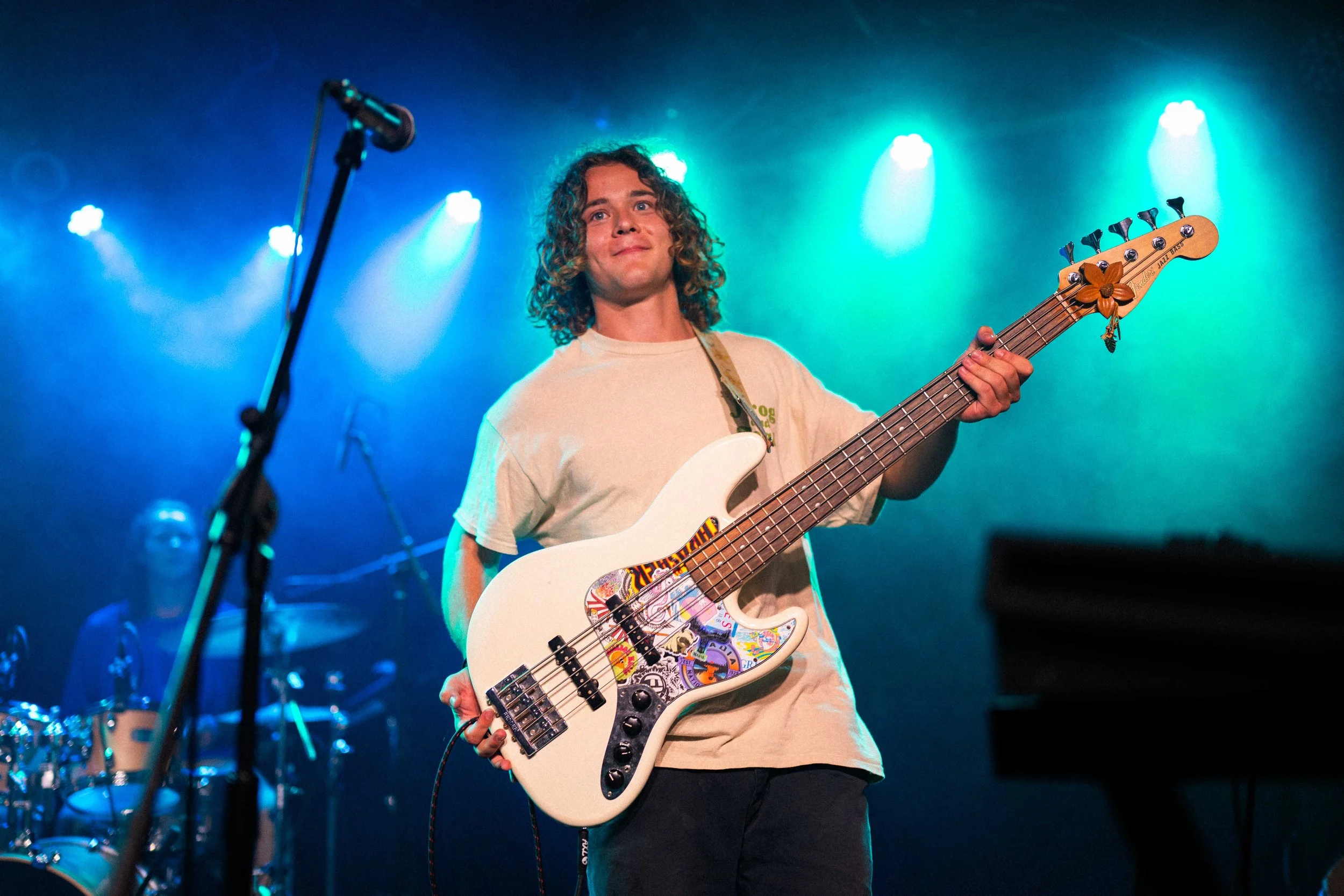 Young man with curly hair on stage holding a white electric bass guitar, colorful stickers on it, under blue and green stage lights, with a drummer in the background and a keyboard on the right.