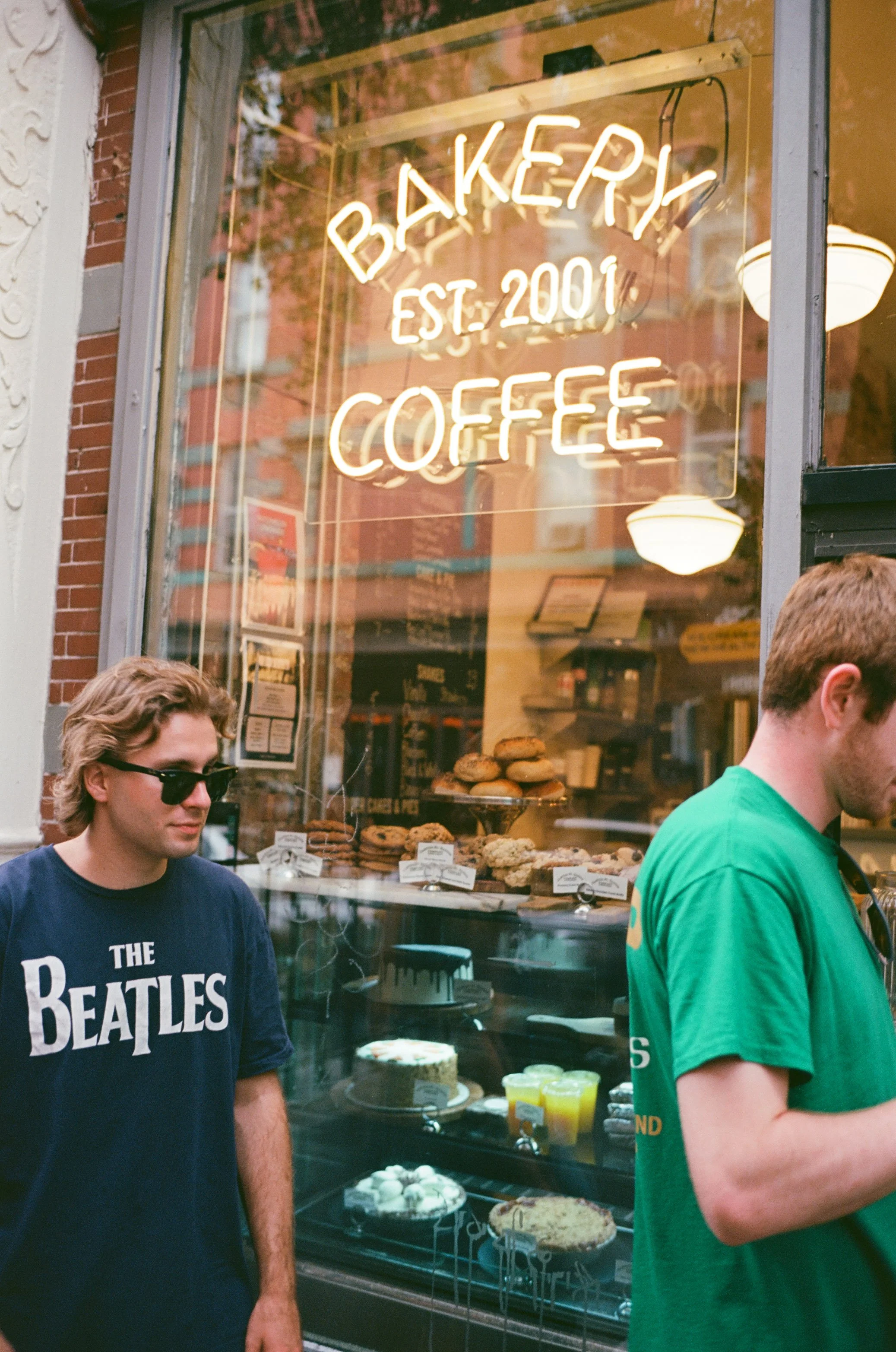A bakery window displaying various baked goods with a neon sign that reads 'Bakery Est. 2001 Coffee.' Two young men stand outside, one wearing sunglasses and a Beatles t-shirt, the other in a green shirt.