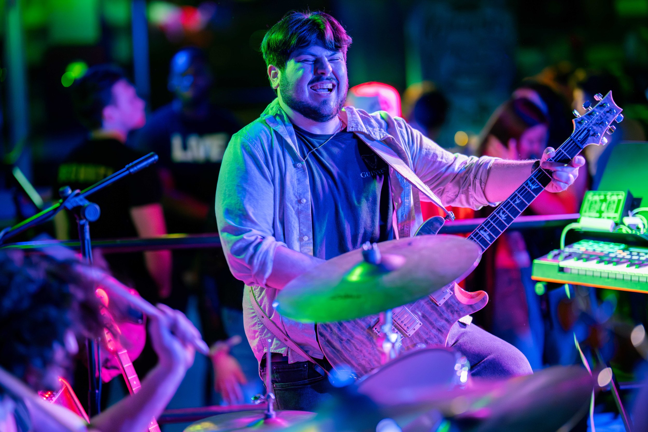 A musician playing the guitar and drums at a concert venue with colorful neon lights and a crowd in the background.