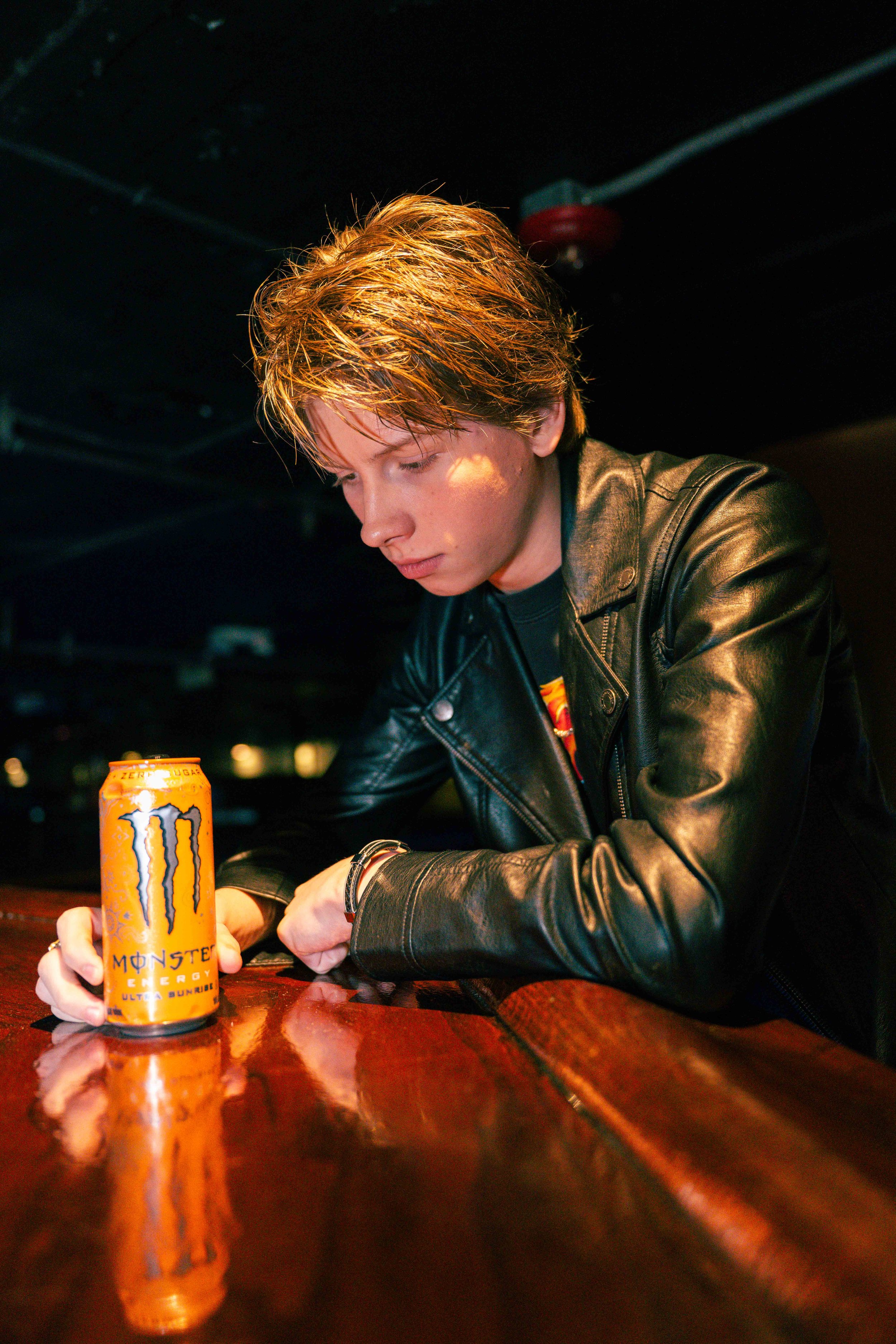 Young man with red hair wearing a black leather jacket sitting at a bar, looking down with a can of Monster Energy drink on the bar in front of him.