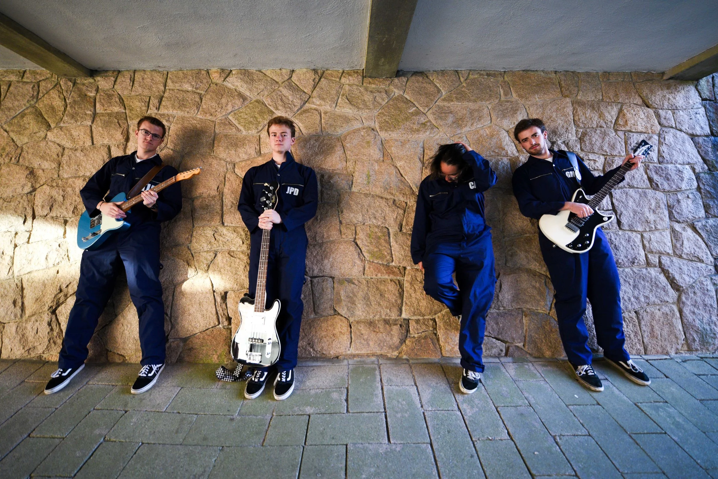 A group of four young people in matching dark uniforms with patches, each holding or standing near a musical instrument, against a stone wall. They have casual poses, with some looking directly at the camera and others looking downward or to the side