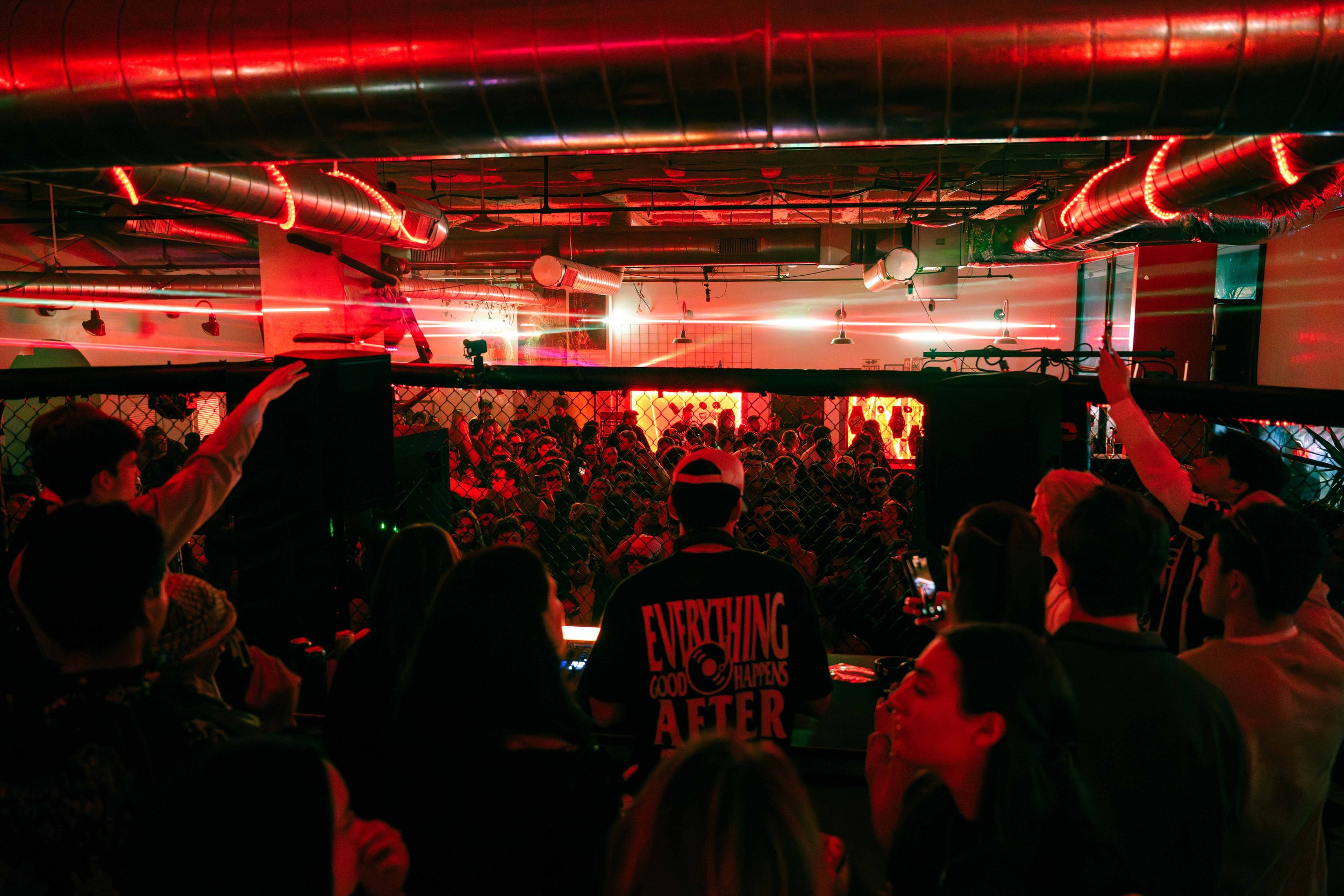 Crowd of people at an indoor event or concert with red and green lighting, industrial-style ceiling, and a DJ in front of the audience.