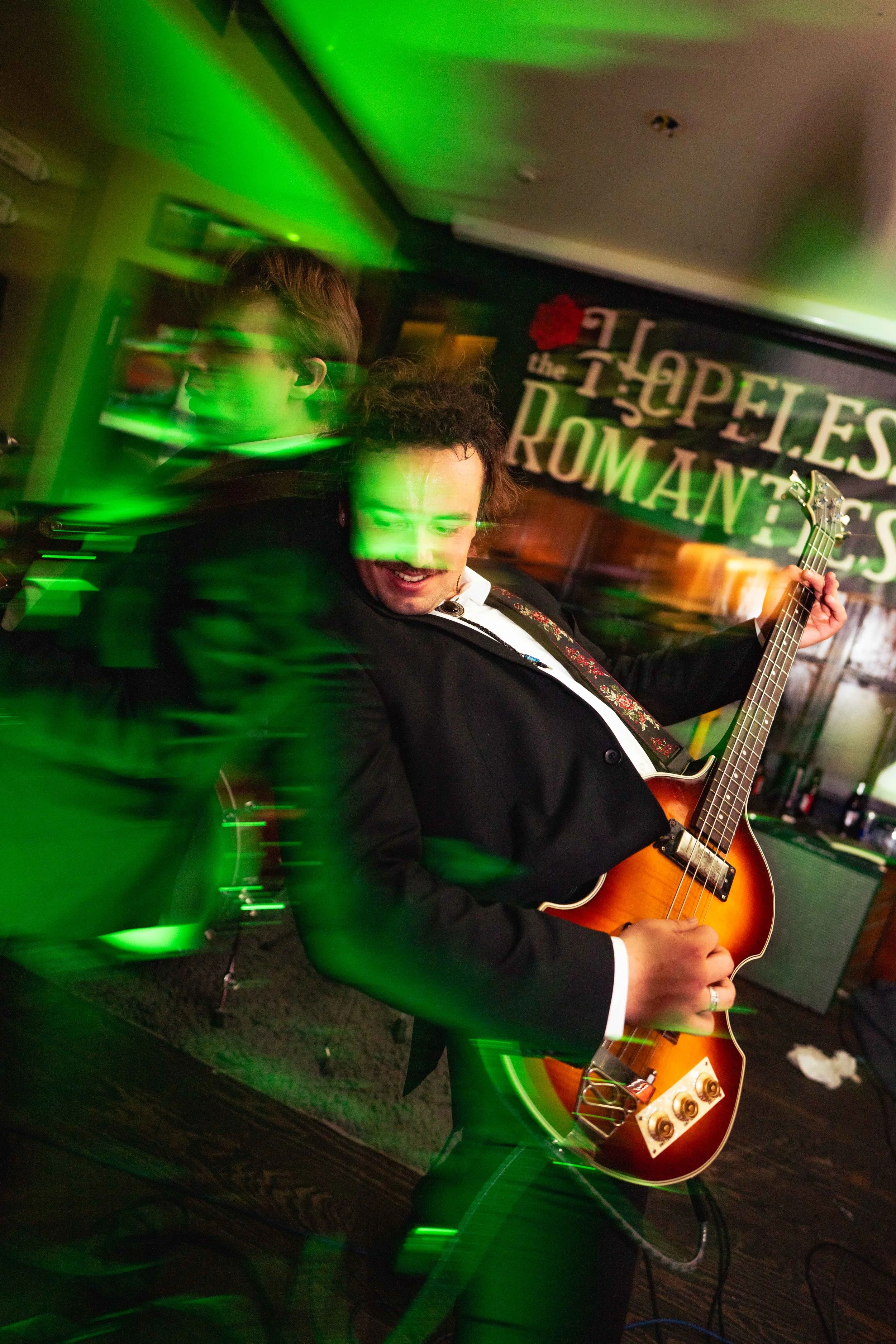 A musician is playing an electric guitar at a bar with green lighting, with a sign in the background that reads 'The Hopeless Romantics'.