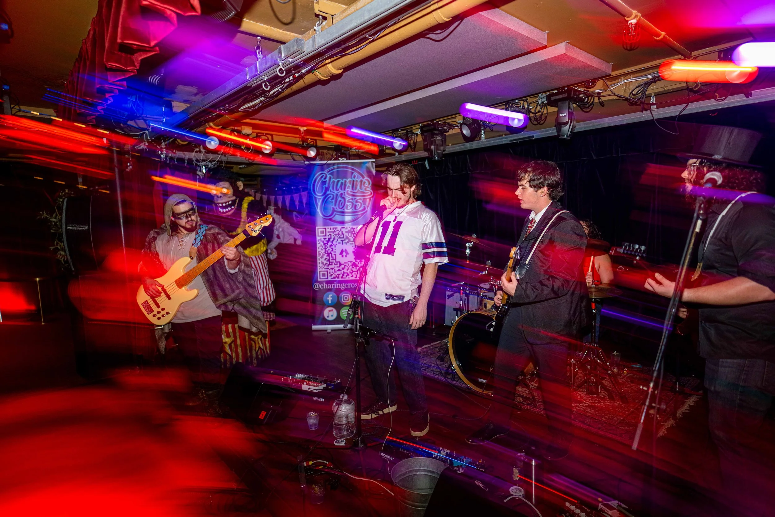 A band performing live on a stage with colorful light streaks, including a guitarist, a bassist, a singer, and a drummer, in a venue with a sign that reads 'Charing Cross' in the background.