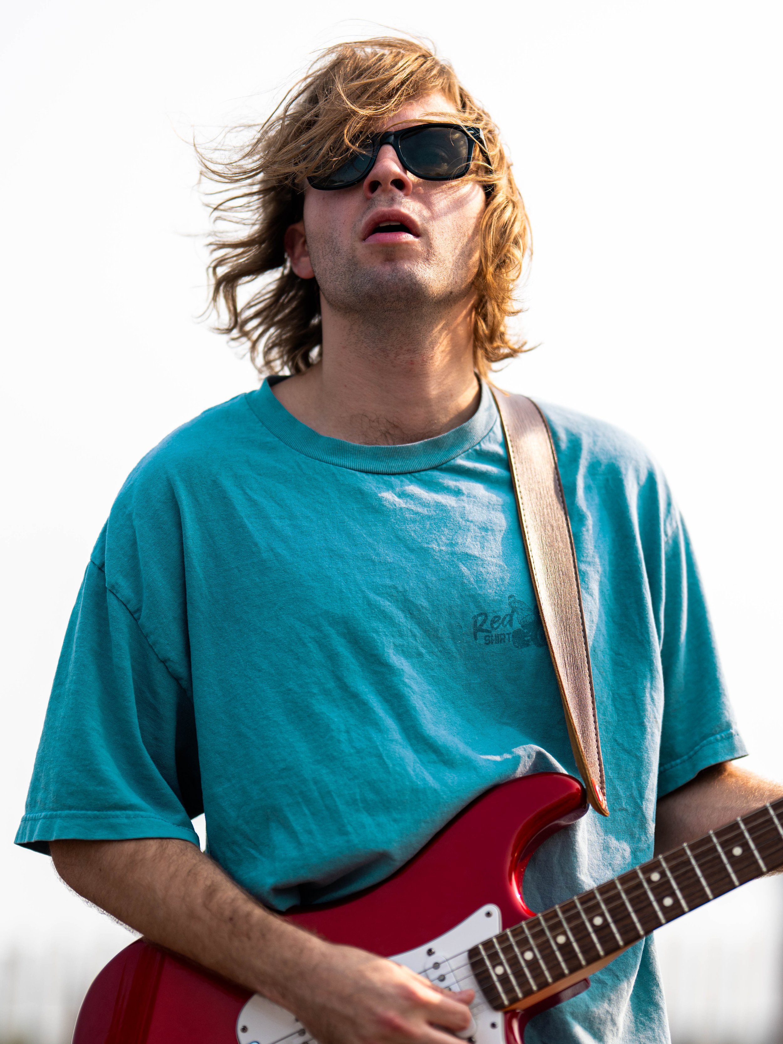 A young man with long, wavy blond hair wearing sunglasses, a light blue T-shirt, and playing a red electric guitar outdoors.