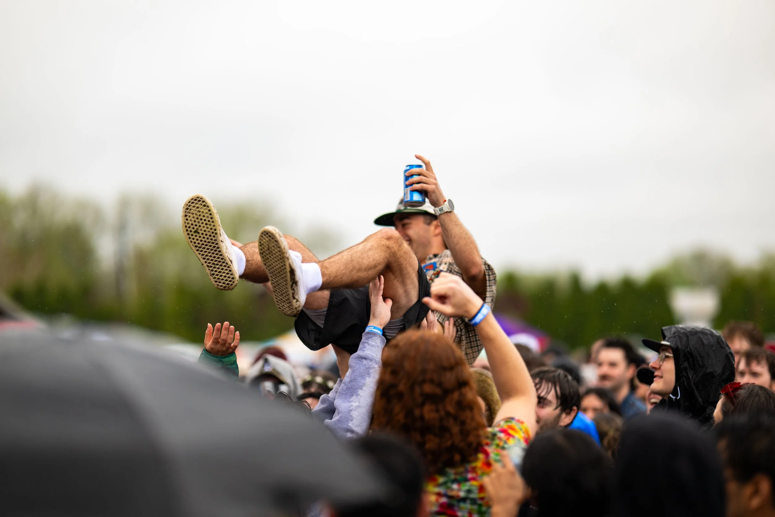 A crowd at an outdoor concert or festival, with a man being lifted above the crowd holding a beverage, smiling, and wearing a hat, surrounded by people in rainy weather.