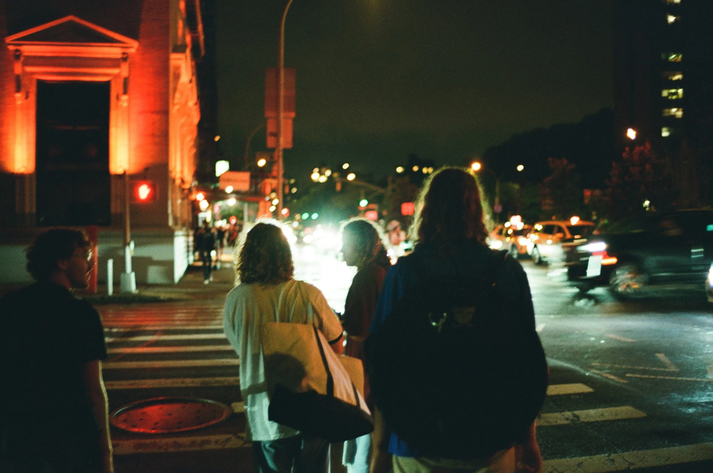 People walking across a busy city street at night with illuminated buildings and cars.
