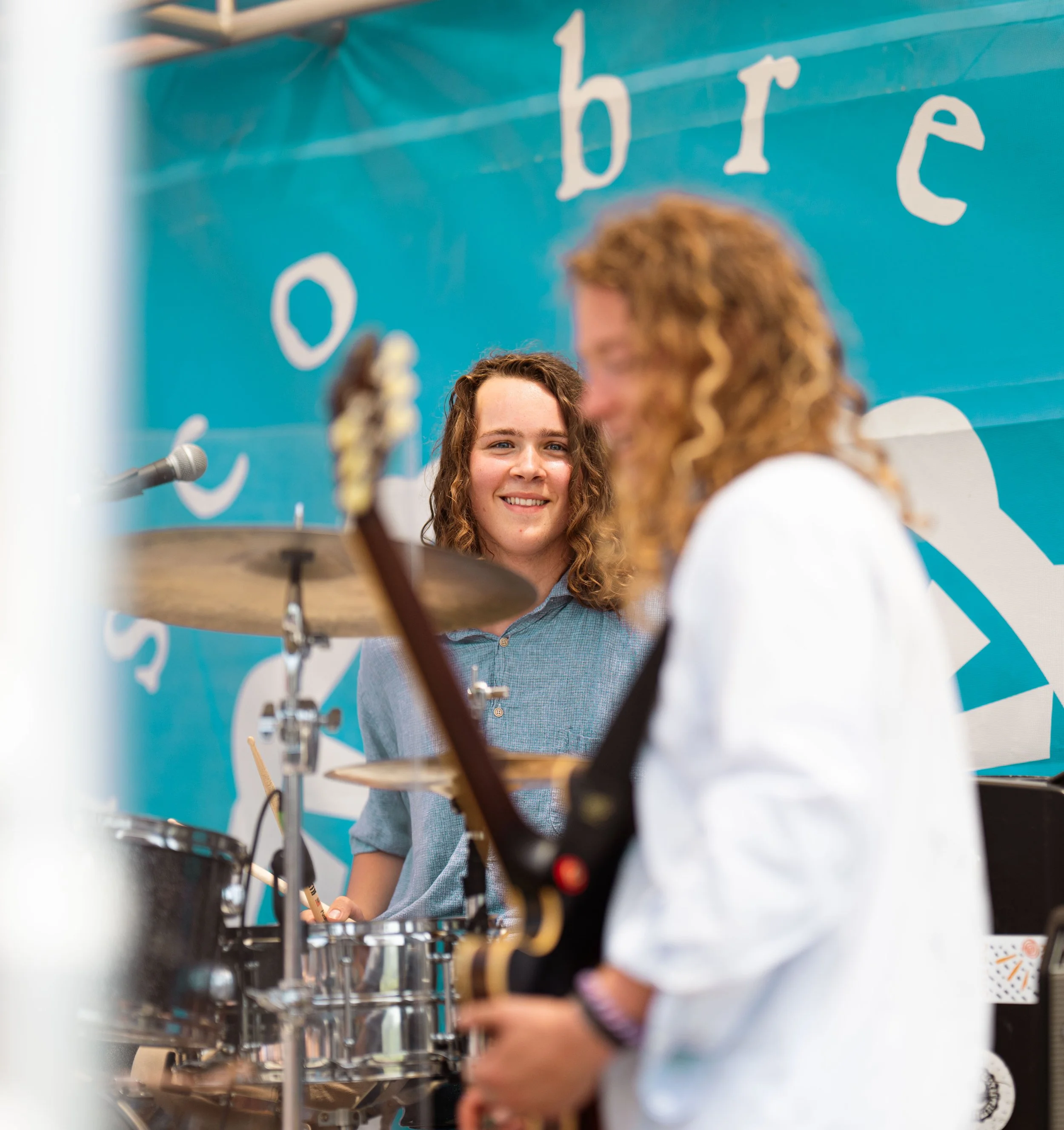 Two young men with long curly hair, one smiling, practicing music together in a room with a blue banner that says 'o b re' in the background.