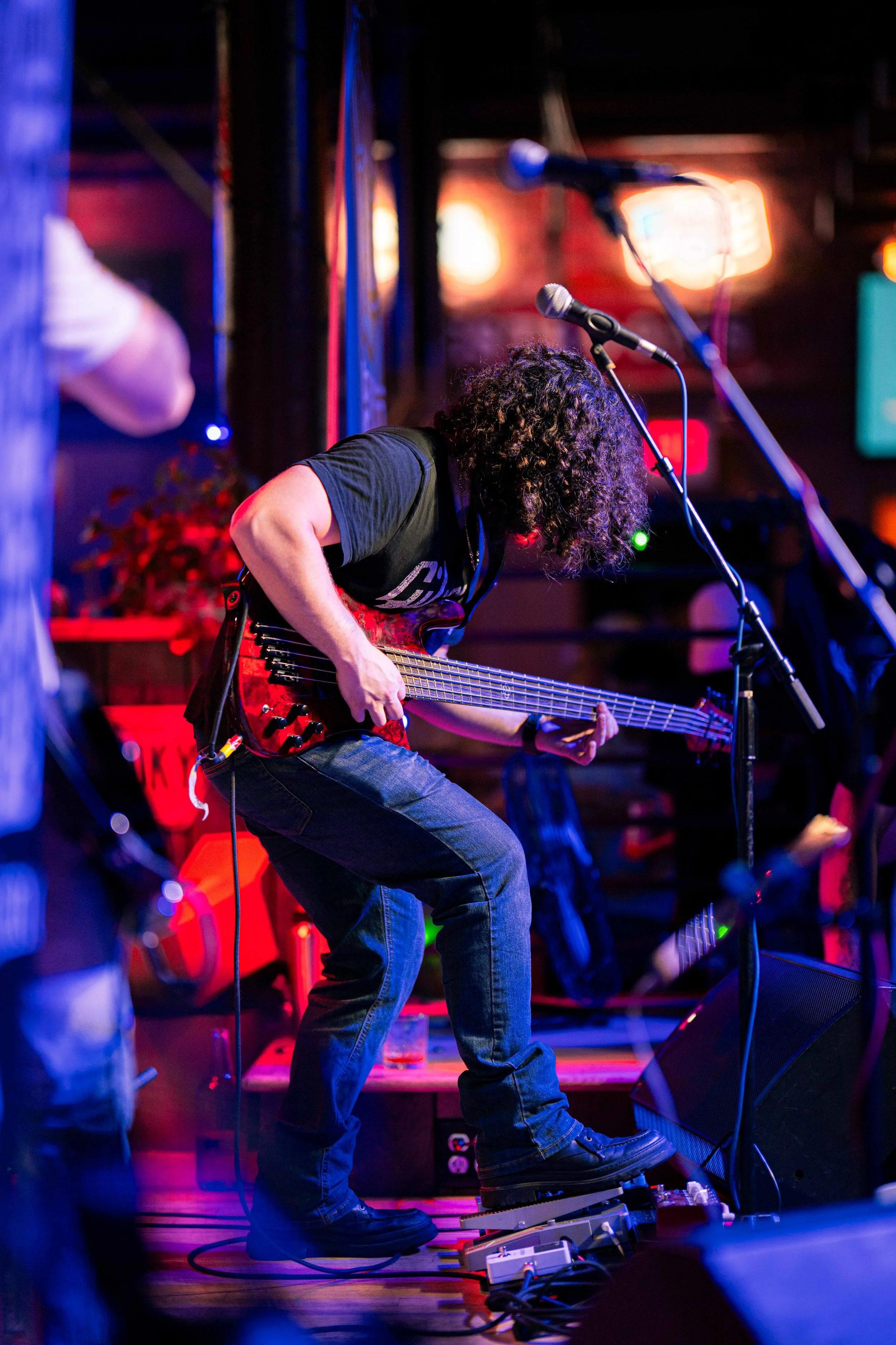 Musician with curly hair playing an electric bass guitar on stage, illuminated by colorful stage lights, with microphones and musical equipment around.