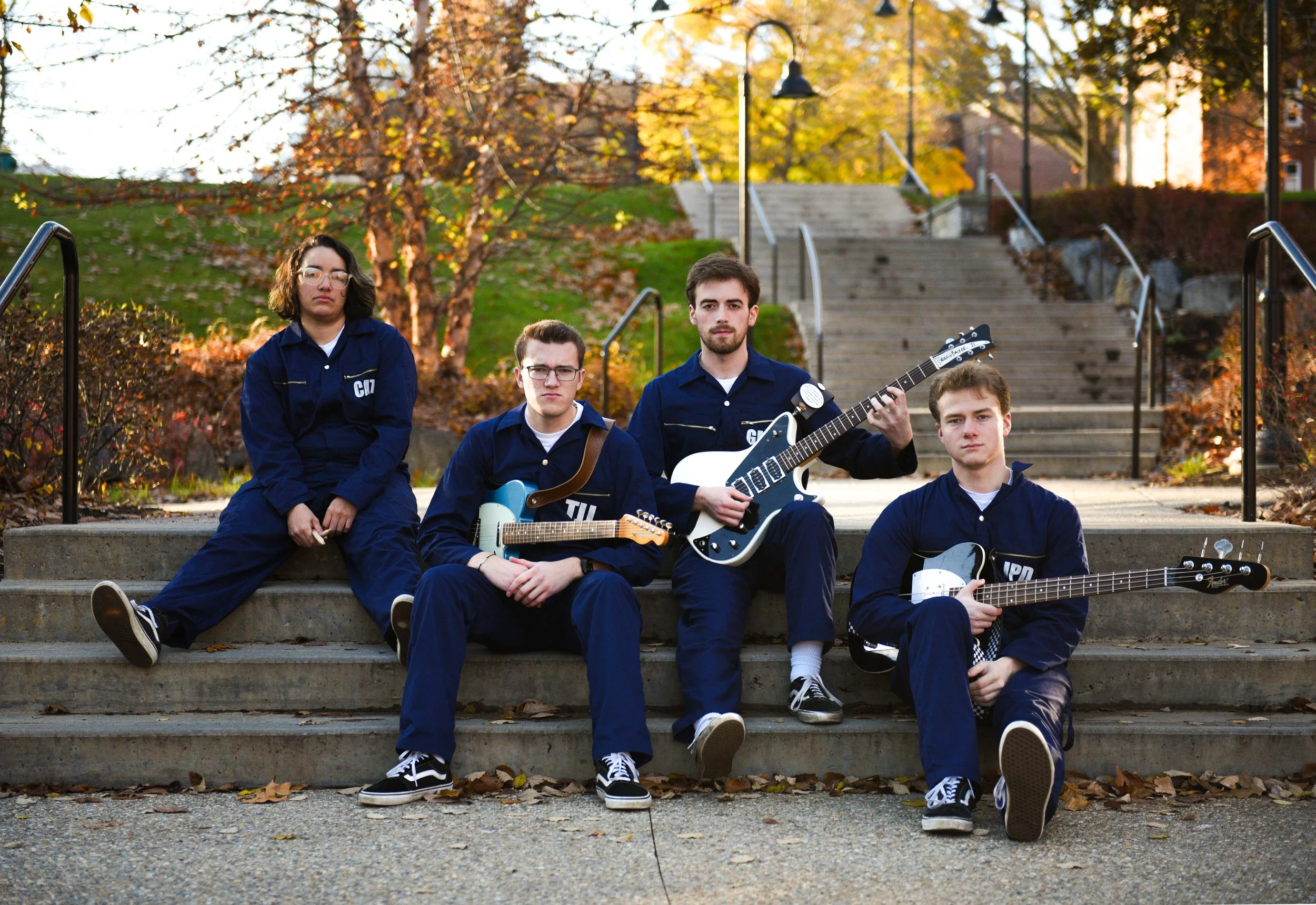 Five young people sitting on stairs outdoors with autumn trees in the background, holding guitars, wearing dark blue uniforms with patches.