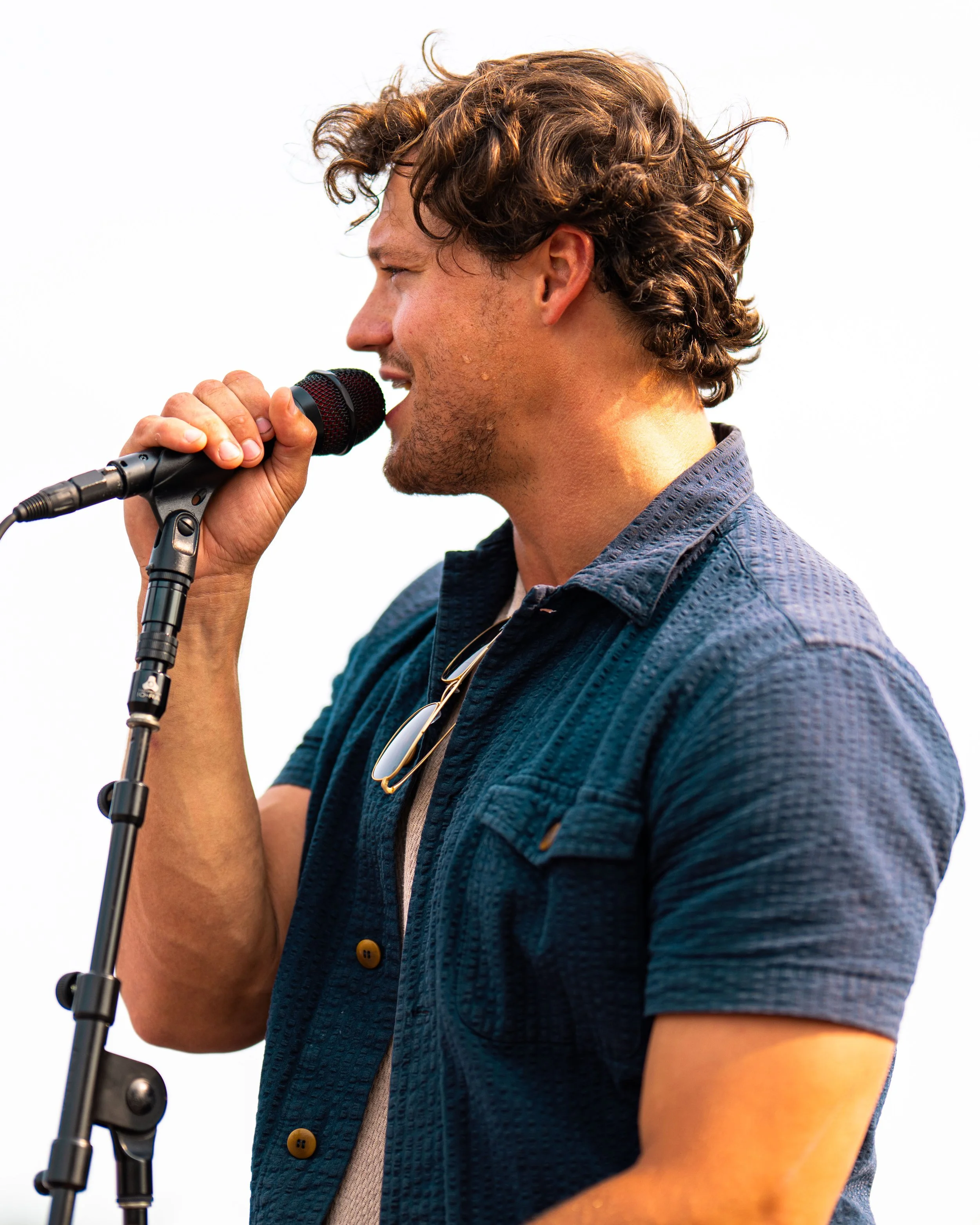 Young man singing into a microphone outdoors, wearing a blue textured shirt and sunglasses hanging from his collar.