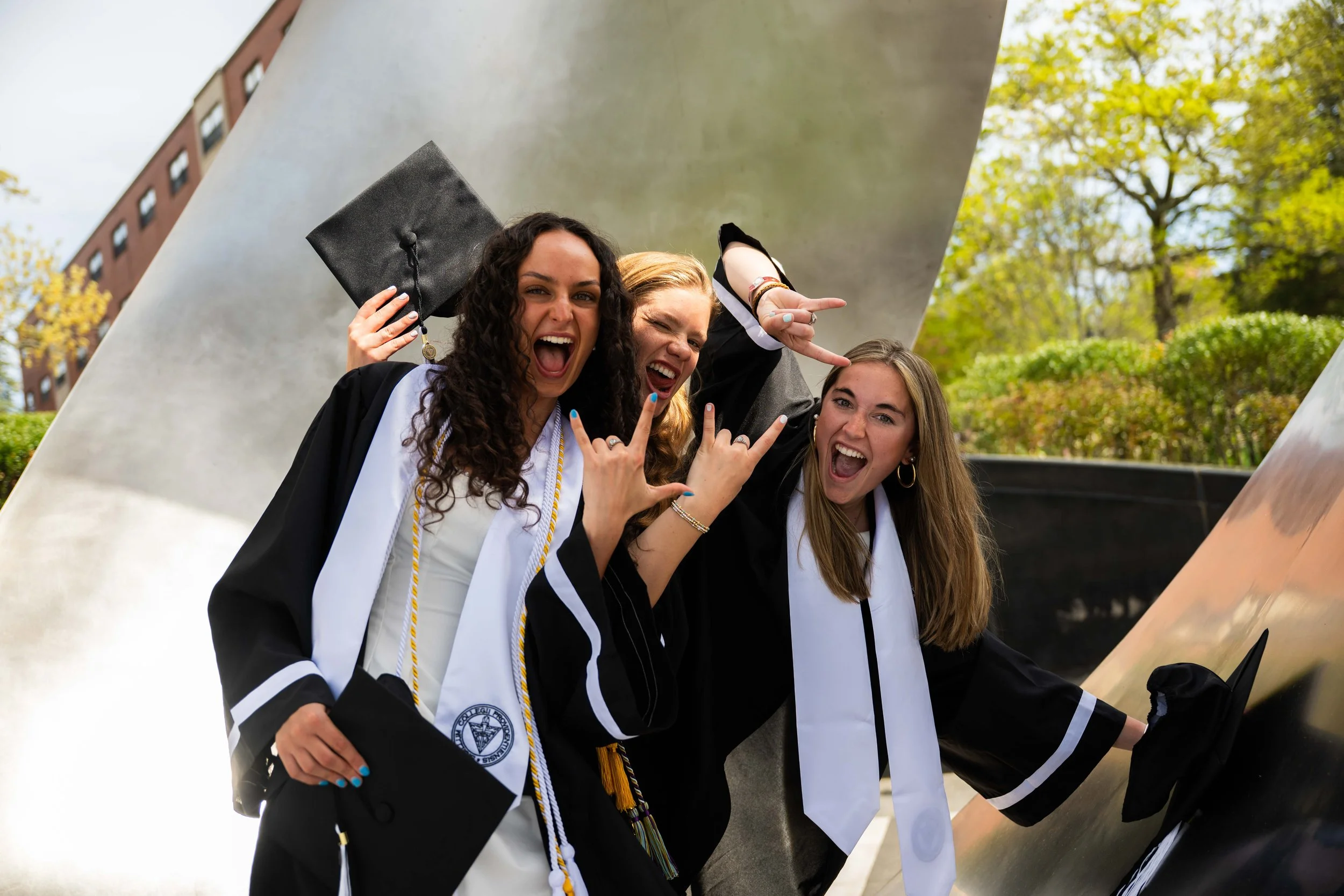Three women in graduation caps and gowns celebrating outdoors, making victory signs and smiling.