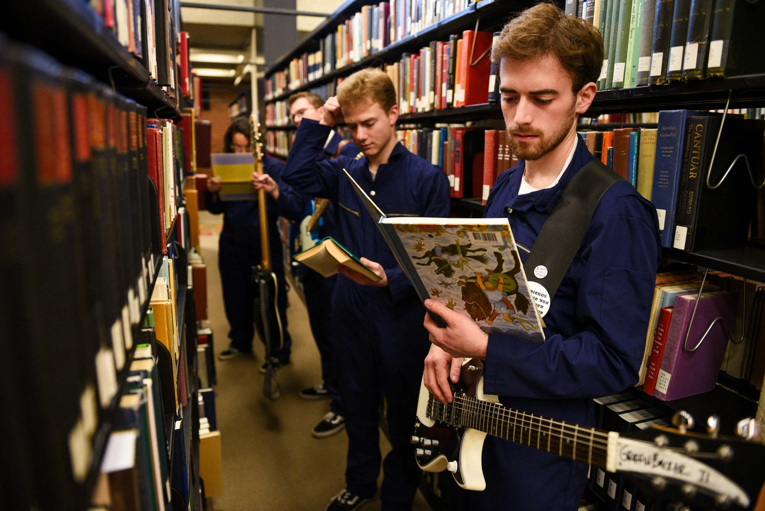 Four young men in navy-blue jackets standing in a library aisle, browsing books, one holding a guitar.
