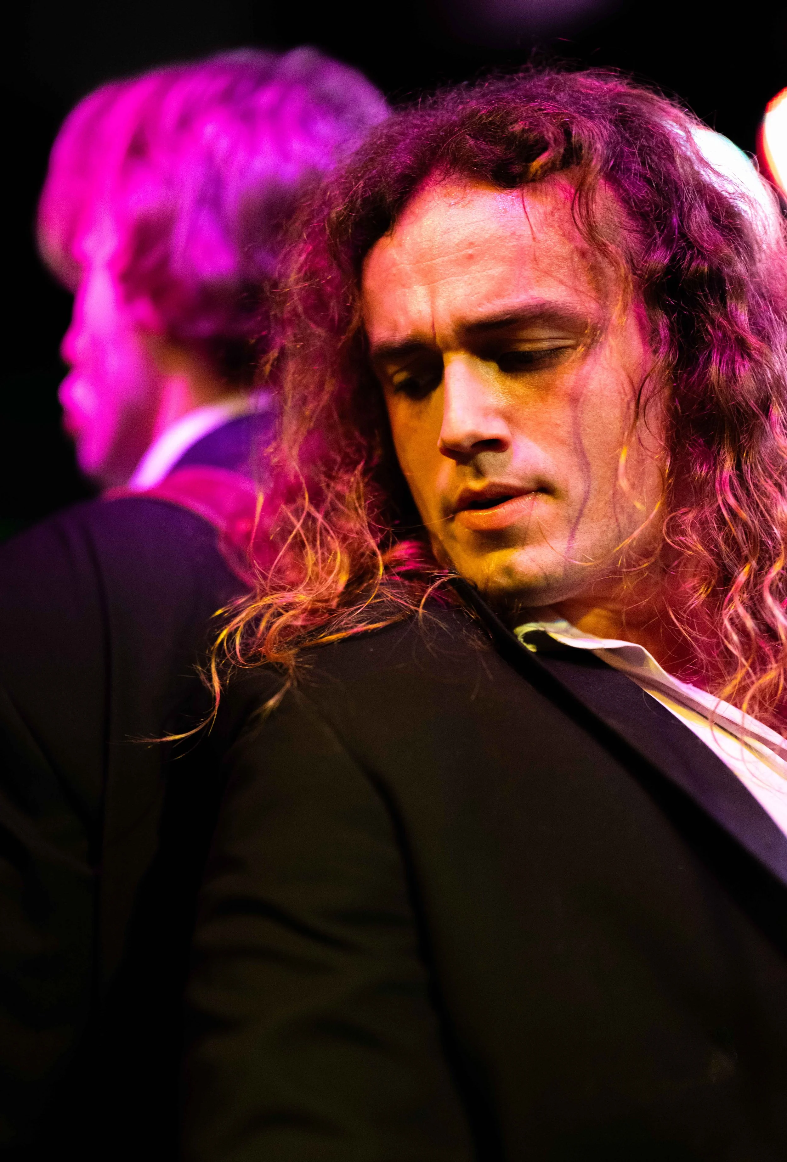 Two men with long hair, one with curly hair in the foreground and the other with straight hair in the background, both dressed in dark suits, in a scene with colorful stage lighting.